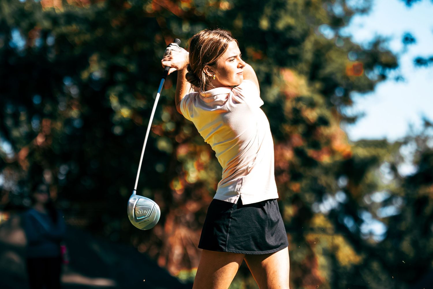 Golfer in white shirt and black skort swings club during autumn round with colorful foliage in background.