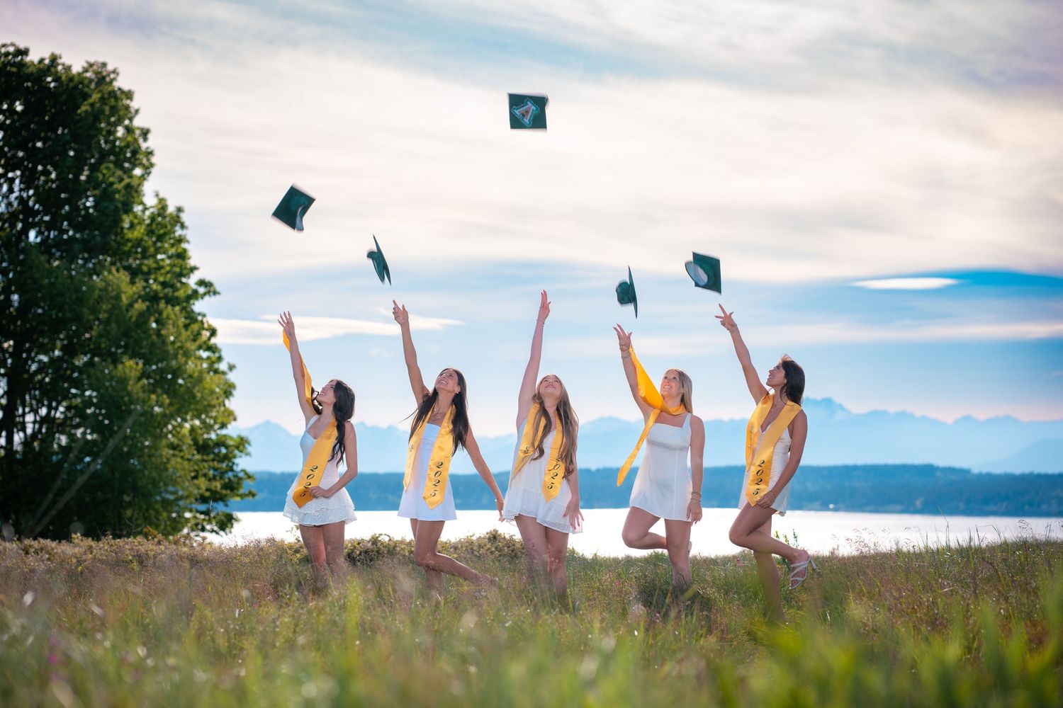 Graduates in white throw their blue graduation caps into the air while standing in a grassy meadow with mountains behind them.