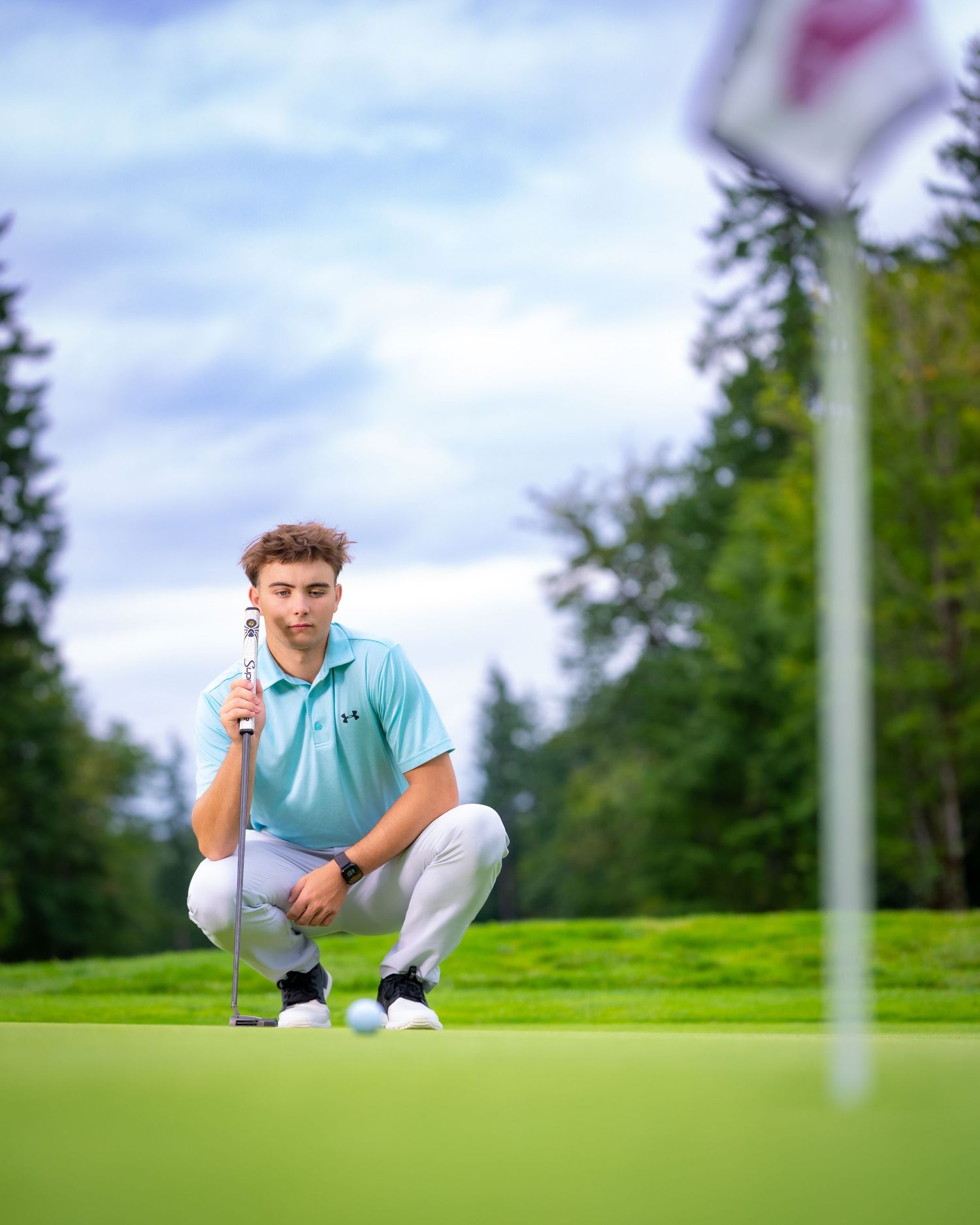 Golfer crouching on green grass to analyze putting line while holding golf ball and club during sunny day.