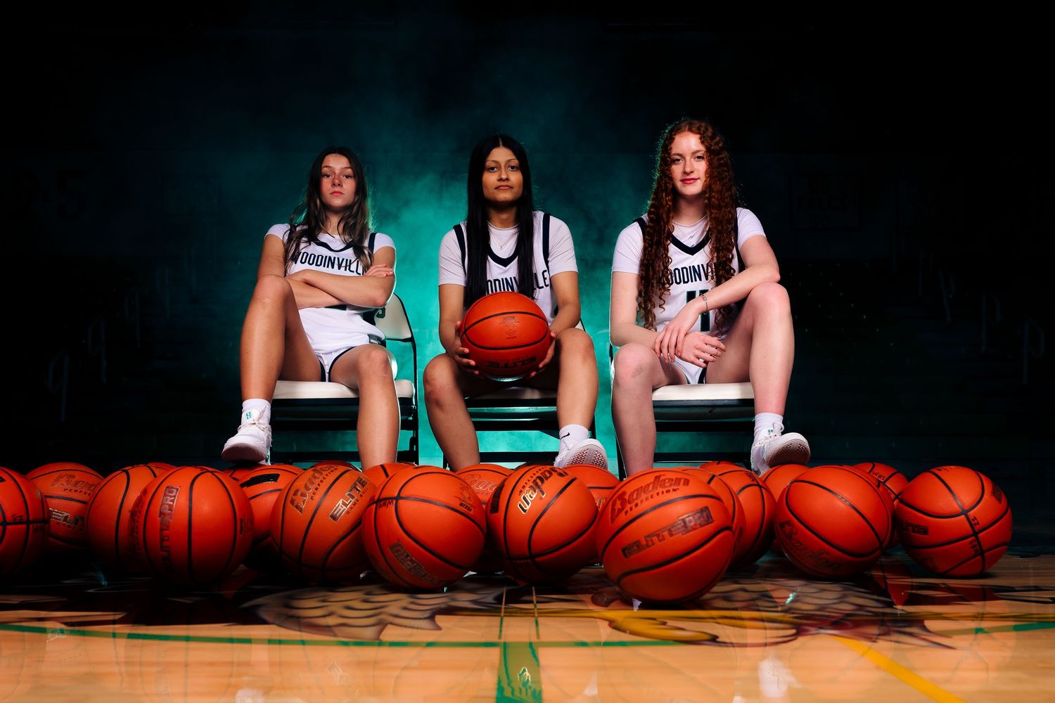 Basketball team photo featuring players sitting with basketballs lined up on wooden court floor.