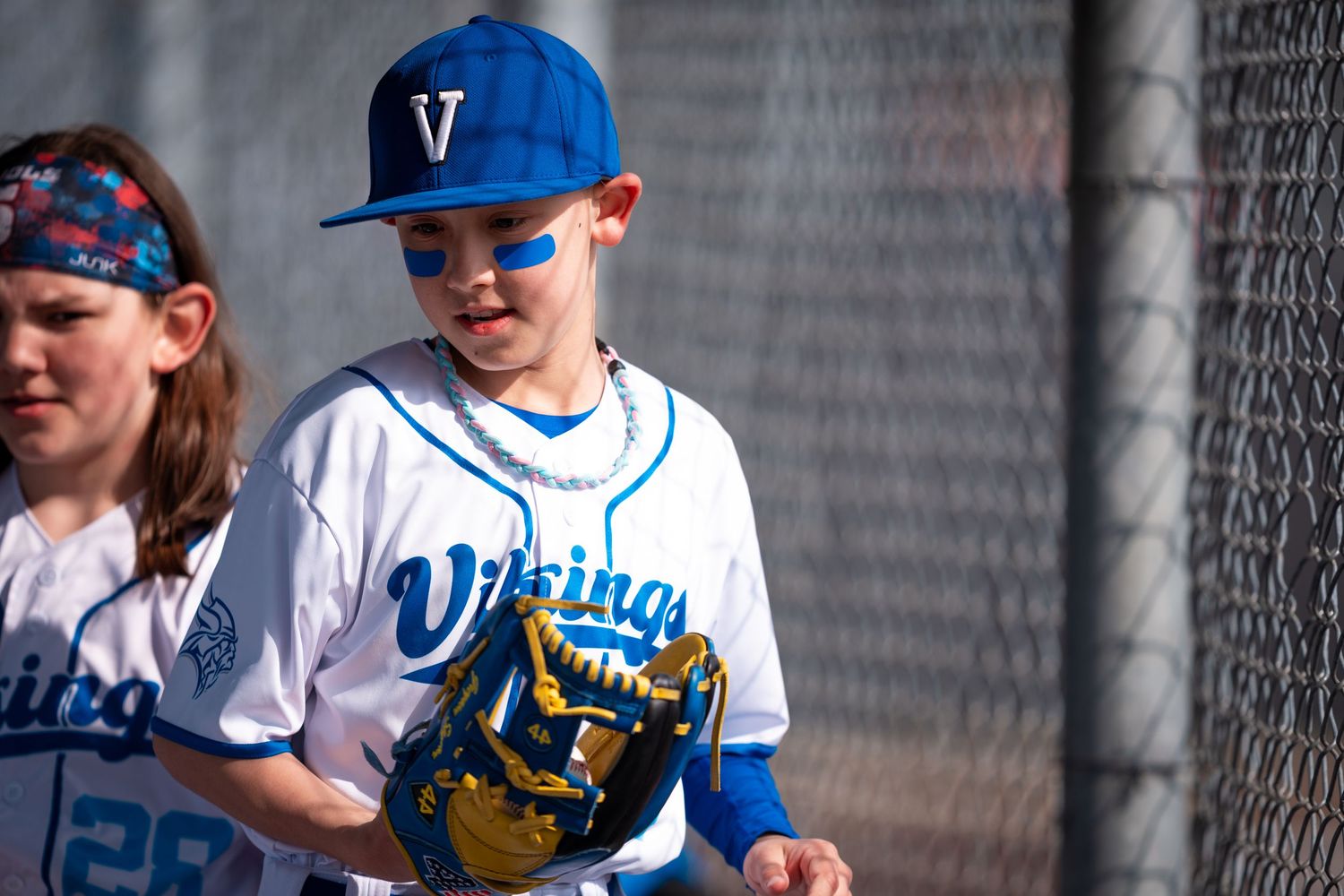 Youth baseball players in blue and white uniforms line up along a chain link fence during a game.