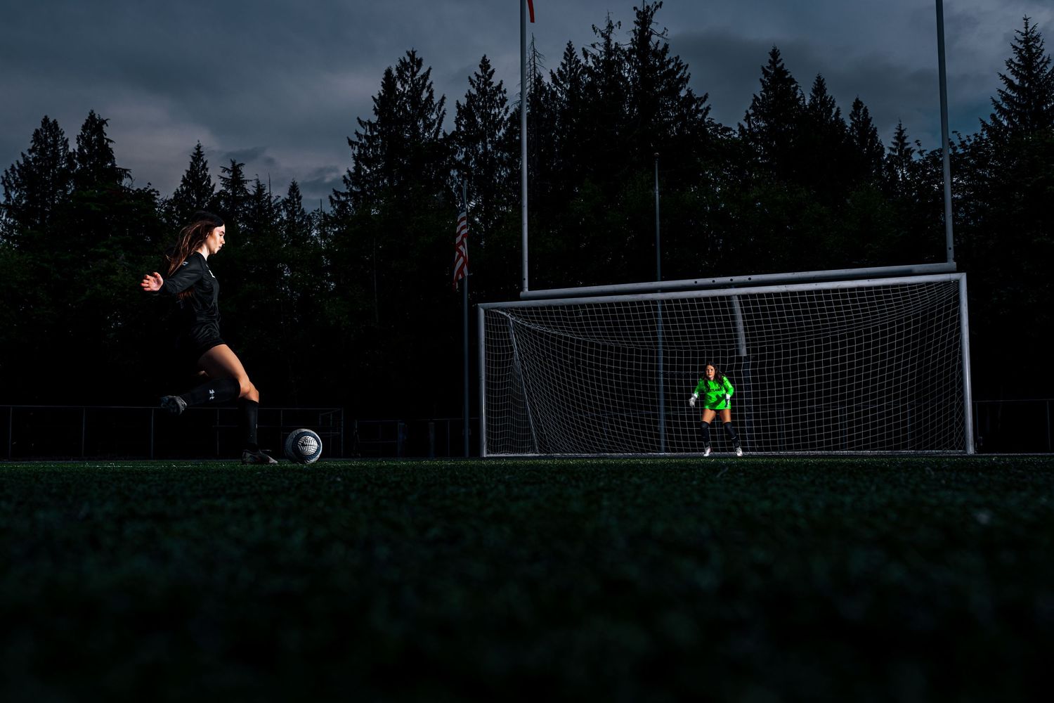 Soccer goalkeeper diving to make a save during a nighttime match at an outdoor field with trees in the background.