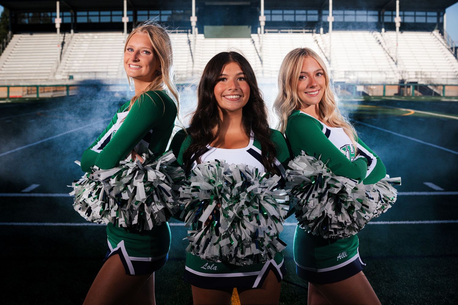 Cheerleaders in green and white uniforms pose with pom poms on a sports field during a sunny day.