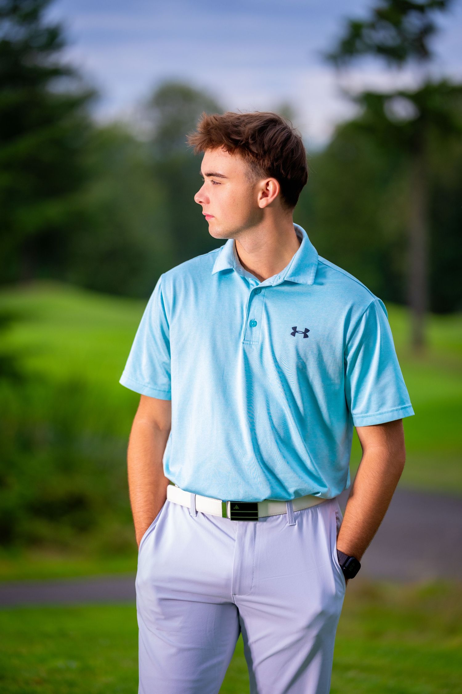 Golfer wearing light blue Under Armour polo and gray pants poses on course.