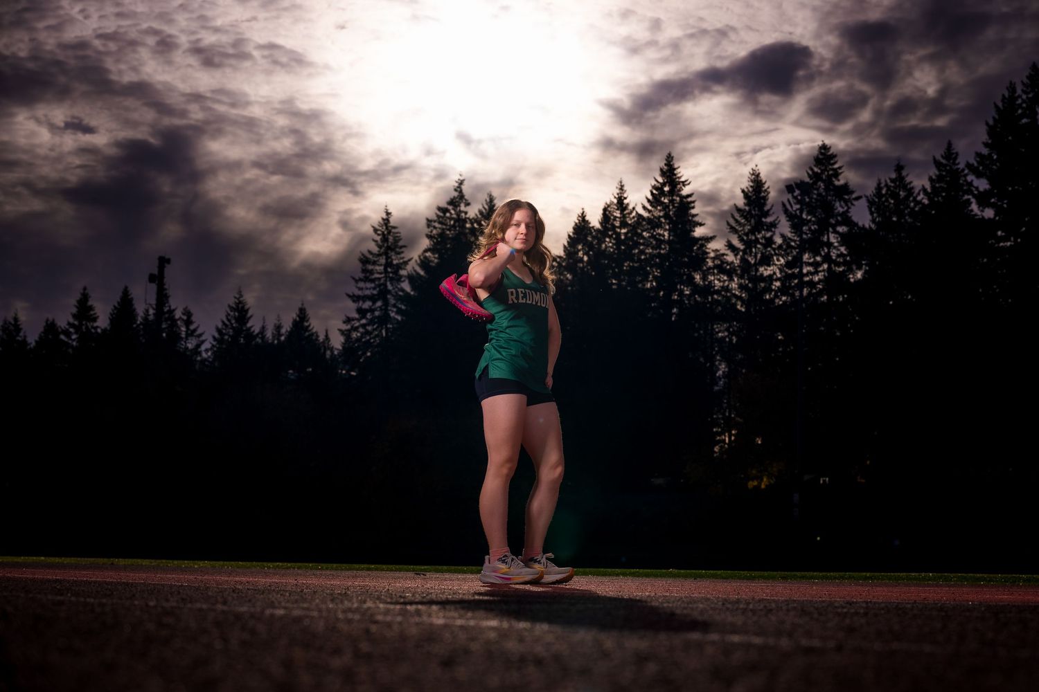 Athletic figure poses in green uniform against dramatic sunset and forest silhouette.