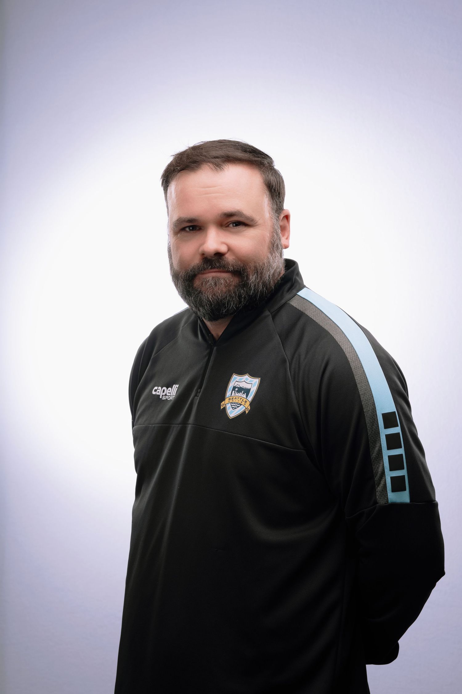 Coach wearing a black polo shirt with team logo poses against white background.
