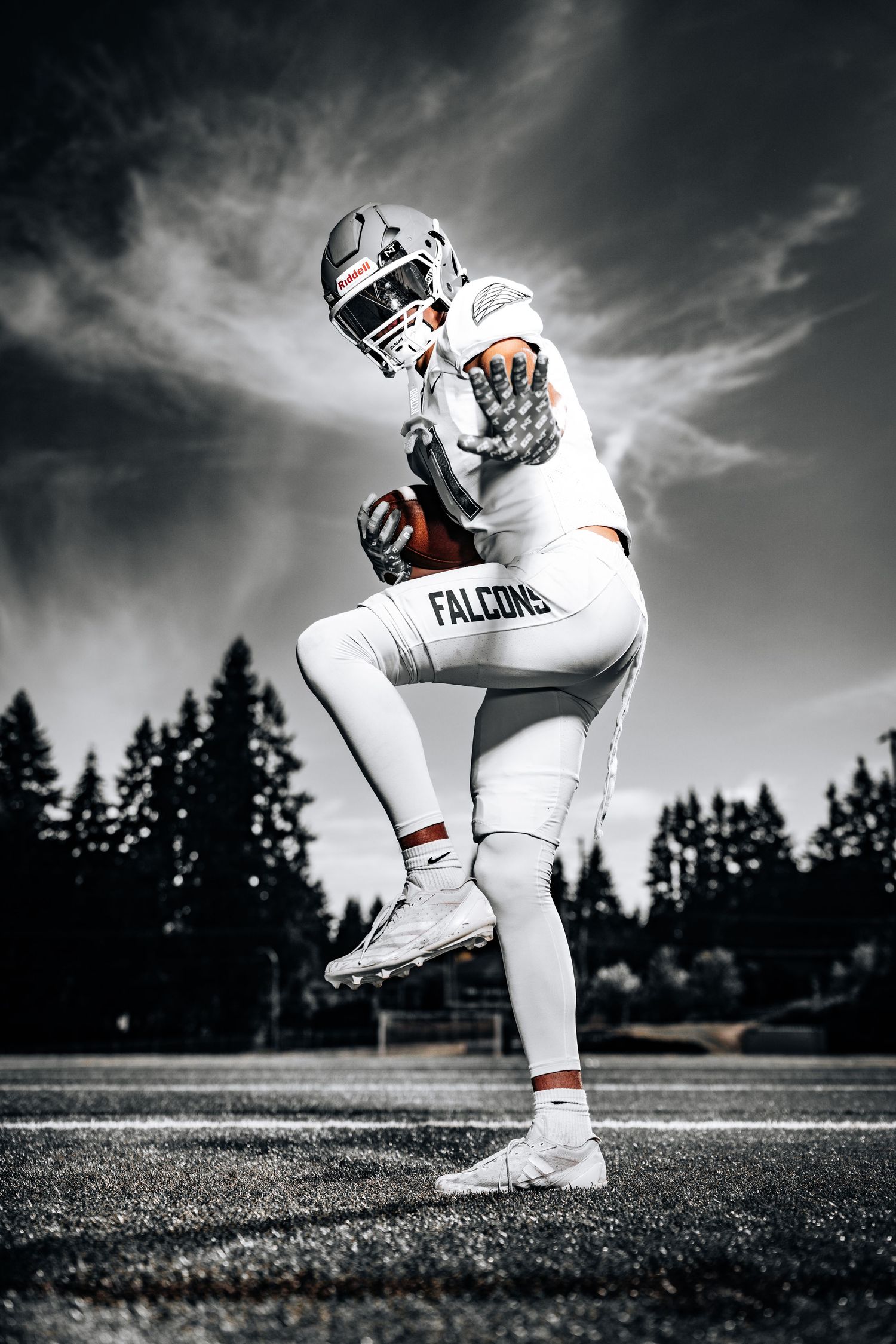Football player in white uniform kneeling on field with dramatic dark sky and trees in background.
