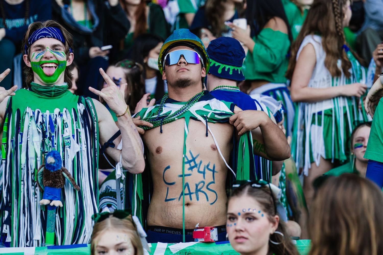 Enthusiastic sports fans in green and blue face paint and tinsel decorations cheer from stadium bleachers.