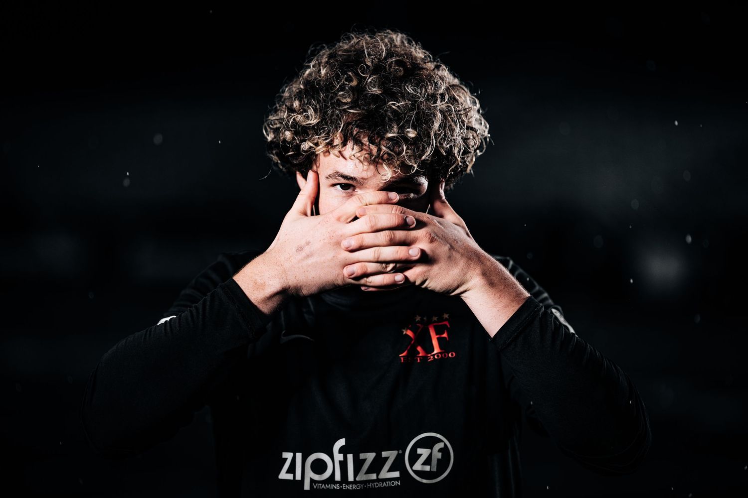 Person in black hoodie covers face with hands against dark background.