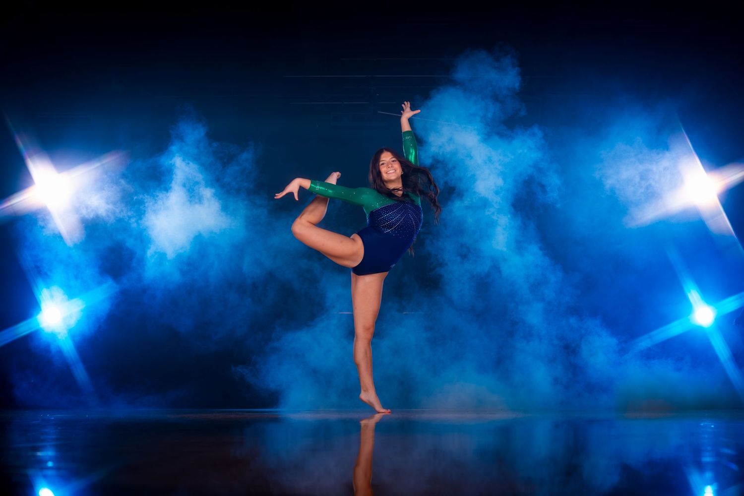 A dancer performs a graceful pose on stage surrounded by blue smoke and glowing star lights in a dramatic setting.