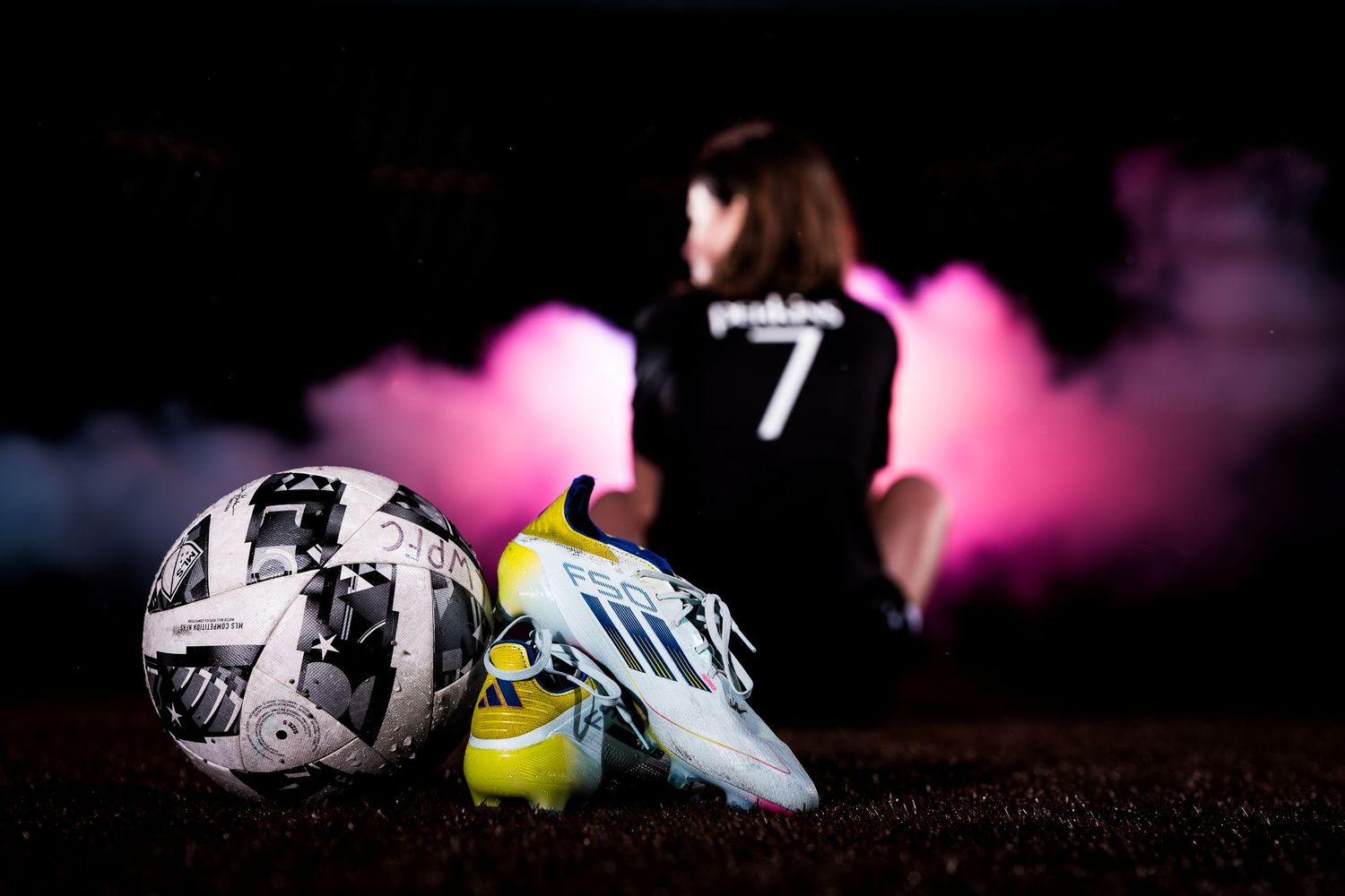 Soccer cleats and sports equipment arranged artistically with pink lighting effects on dark background.