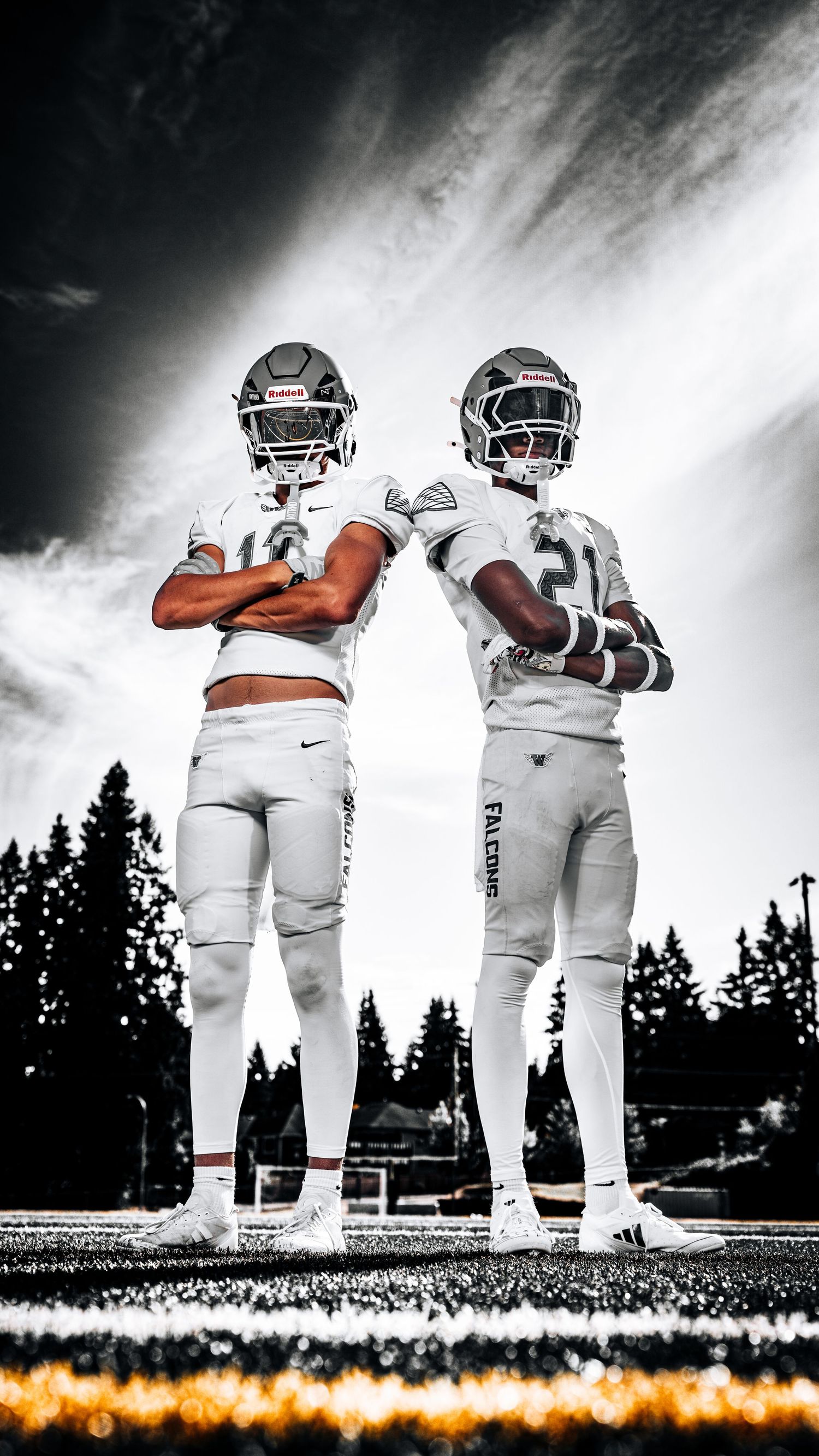 Two football players in white uniforms stand on a field at sunset with dramatic lighting and silhouetted trees in background.