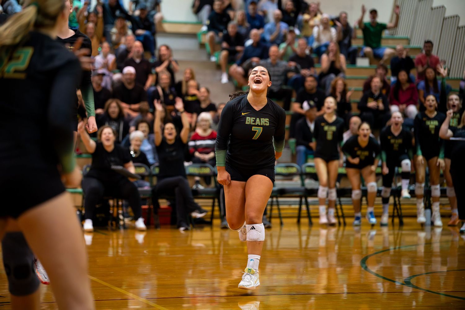 A volleyball player in a black uniform celebrates on the court during a match with spectators in the background.