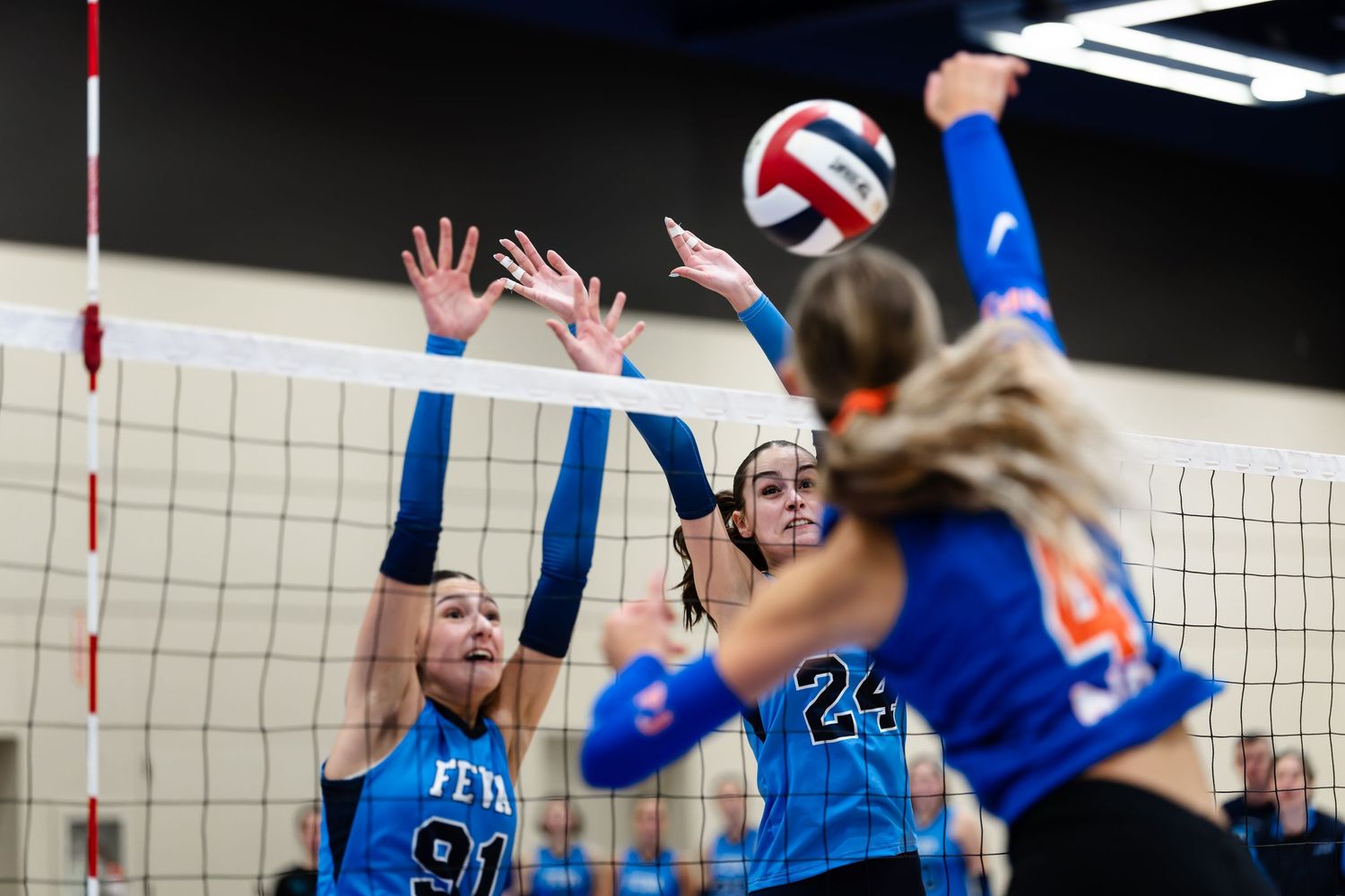 Volleyball players in blue uniforms leap to block a spike at the net during an intense match.