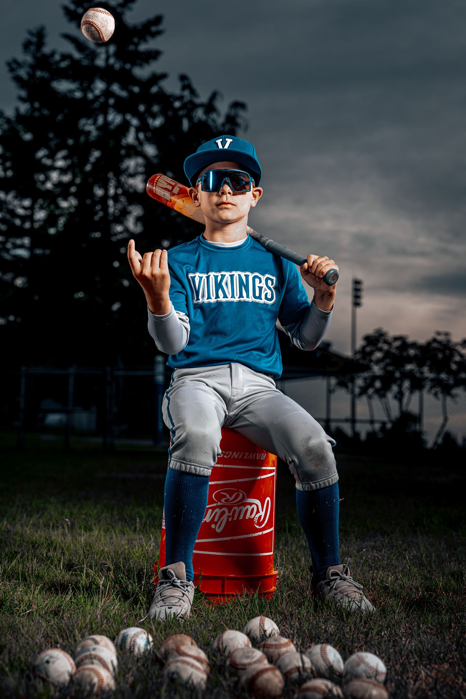 Youth baseball player in Vikings uniform sits on red bucket with baseballs scattered at feet during dramatic sunset.