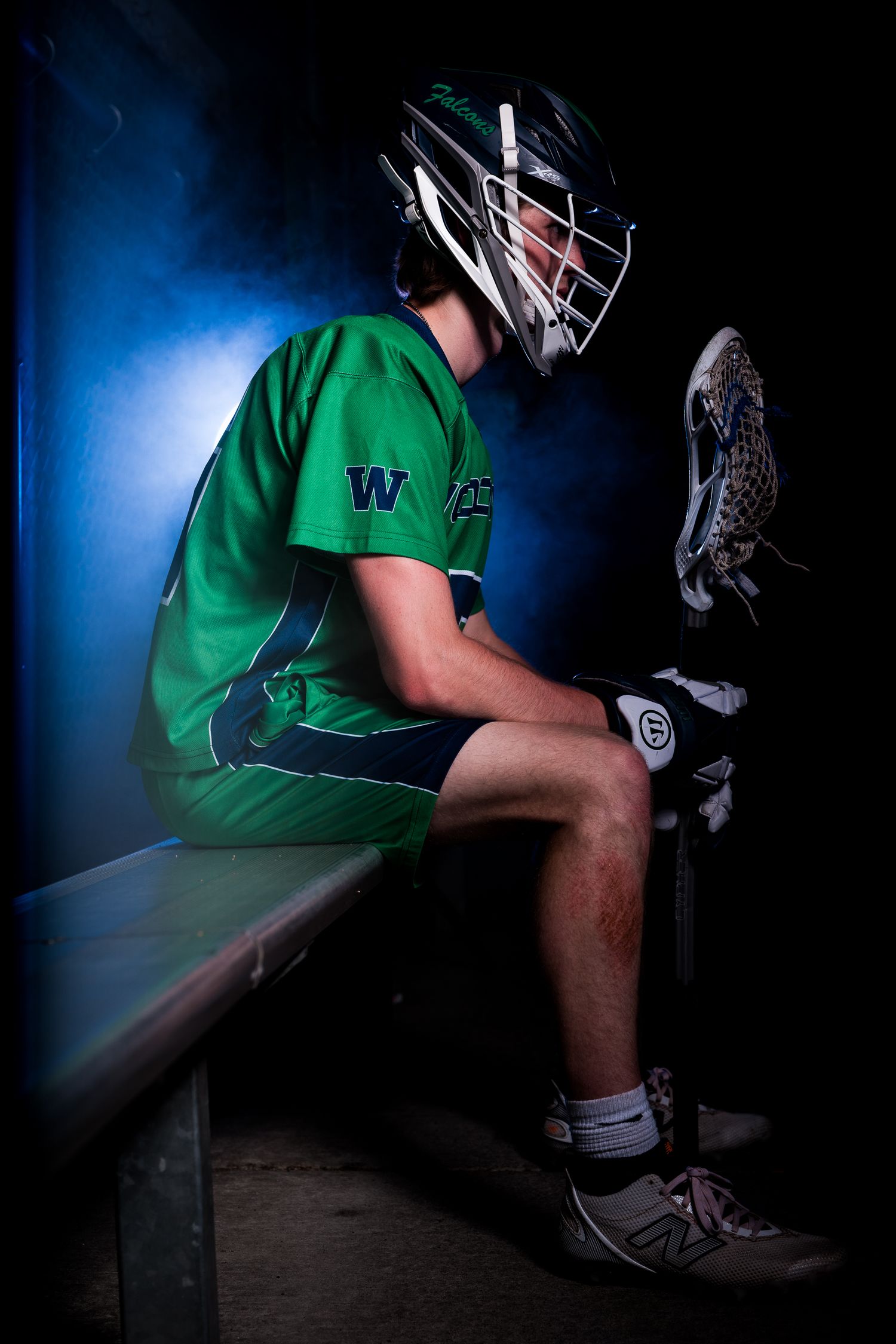 Baseball player in green uniform and helmet sitting in dark dugout with dramatic blue lighting.