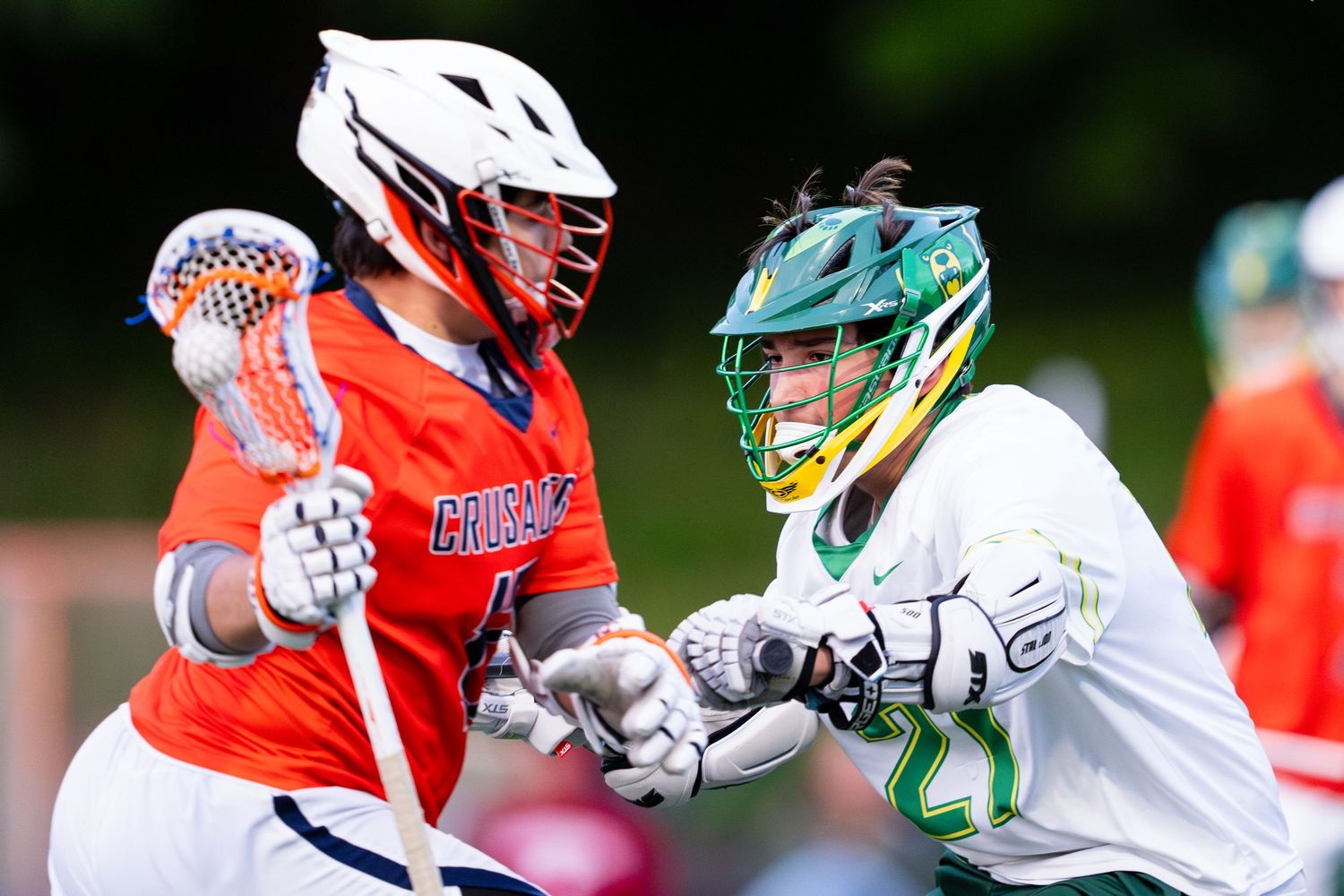 Two lacrosse players in orange and white uniforms face off during an intense match on the field.