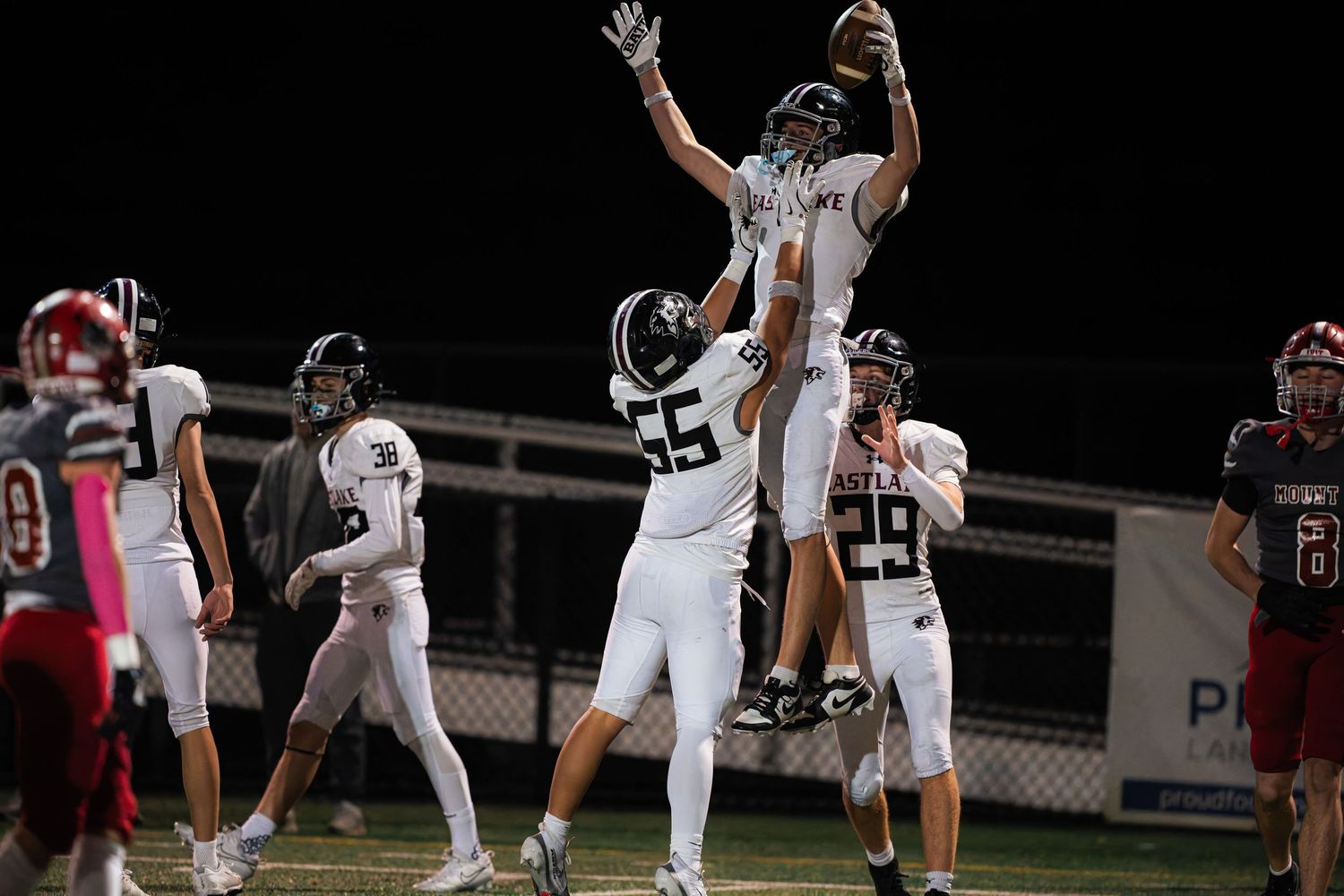 High school football players leap for a catch during a nighttime game under stadium lights.