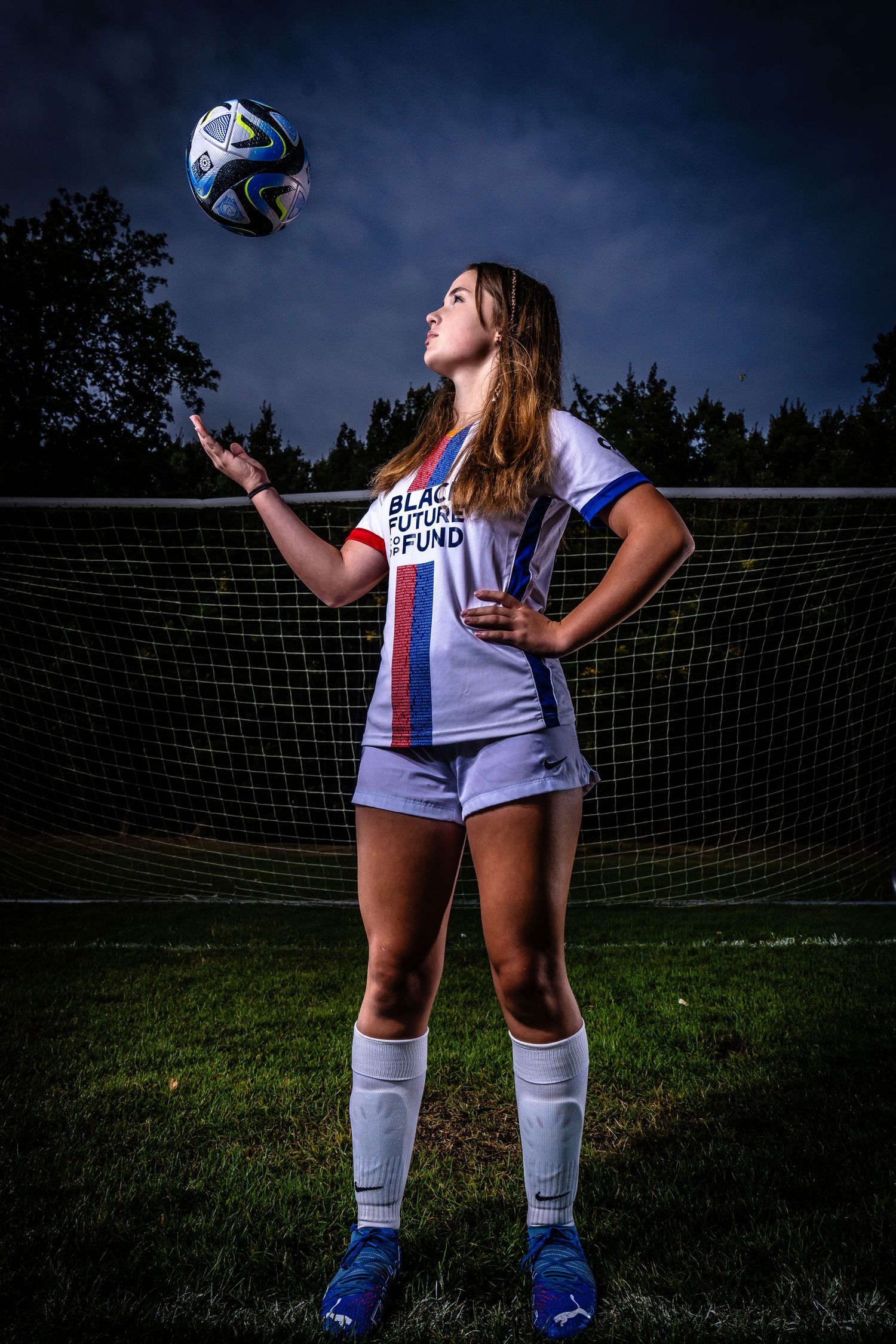 Soccer player in white uniform poses dramatically with ball against dark sky and soccer goal at dusk.