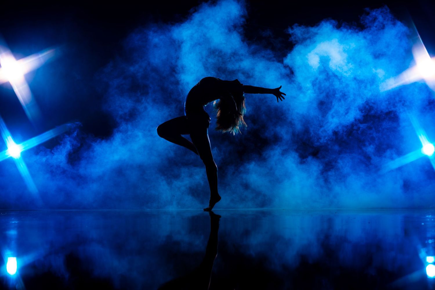 Silhouetted dancer performs graceful pose against dramatic blue misty backdrop with glowing white lights.