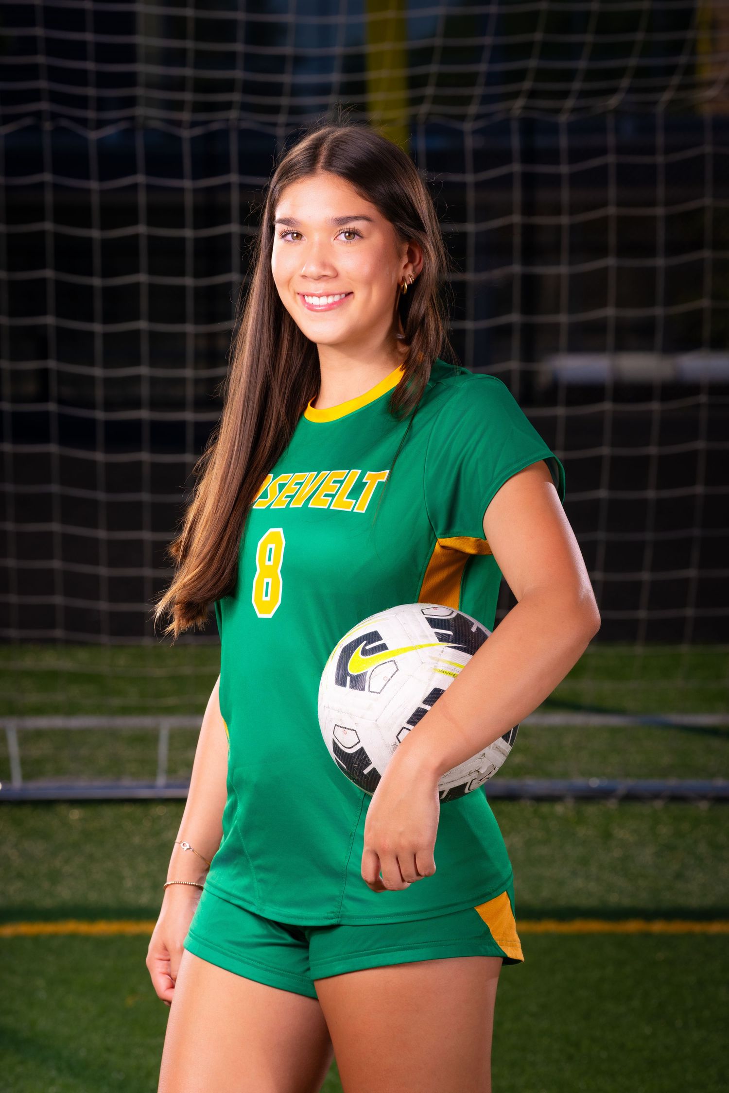 Soccer player in green jersey posing with ball in front of goal net during golden hour.