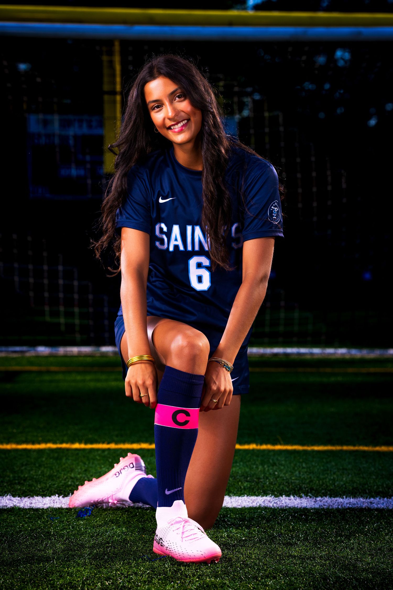 Soccer player in navy blue Saints uniform sits on field wearing pink cleats.