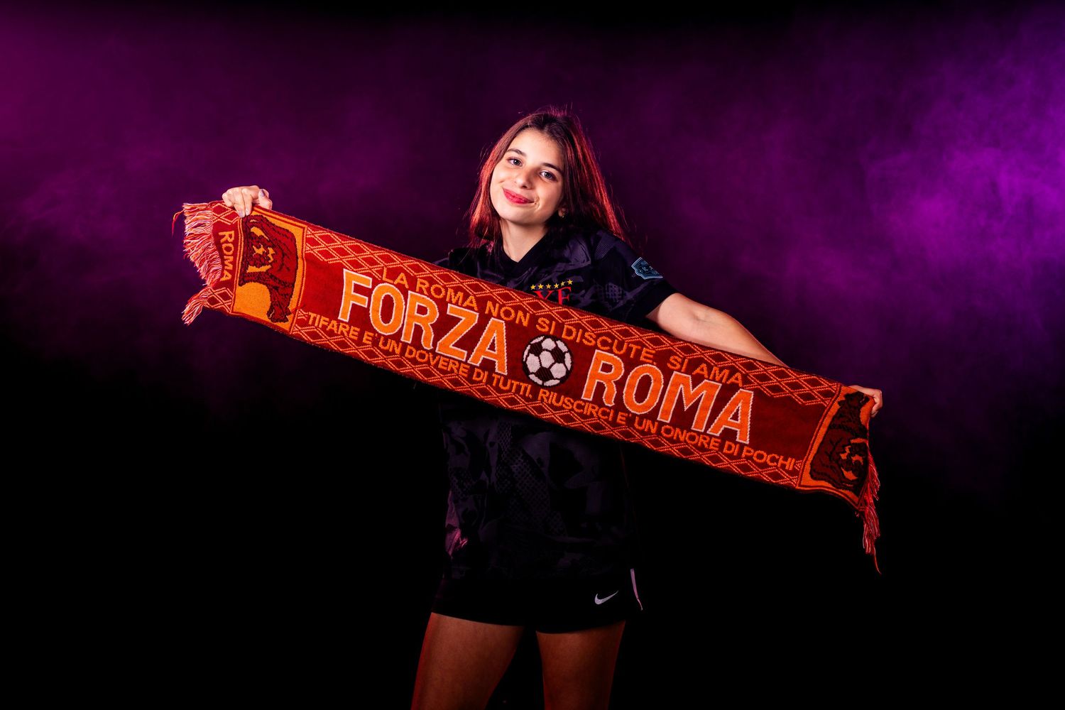 Soccer fans wave red and black team scarves in a dark stadium with purple lighting effects.
