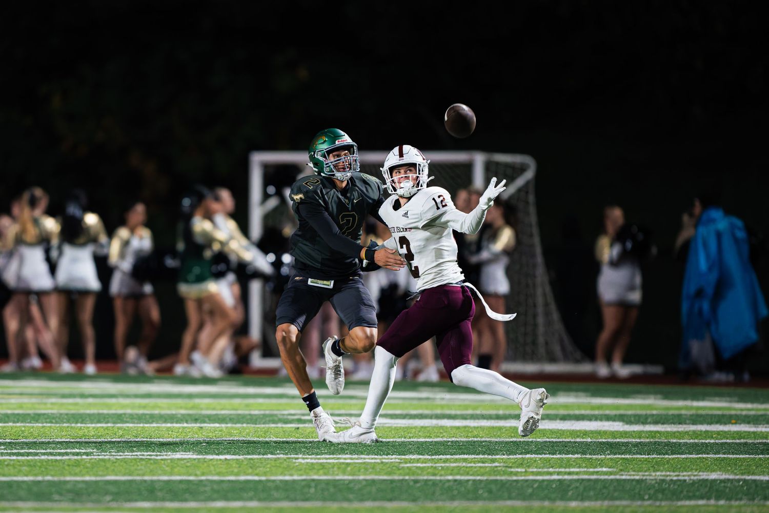 Football player dives to make a catch while another player defends during a night game under stadium lights.