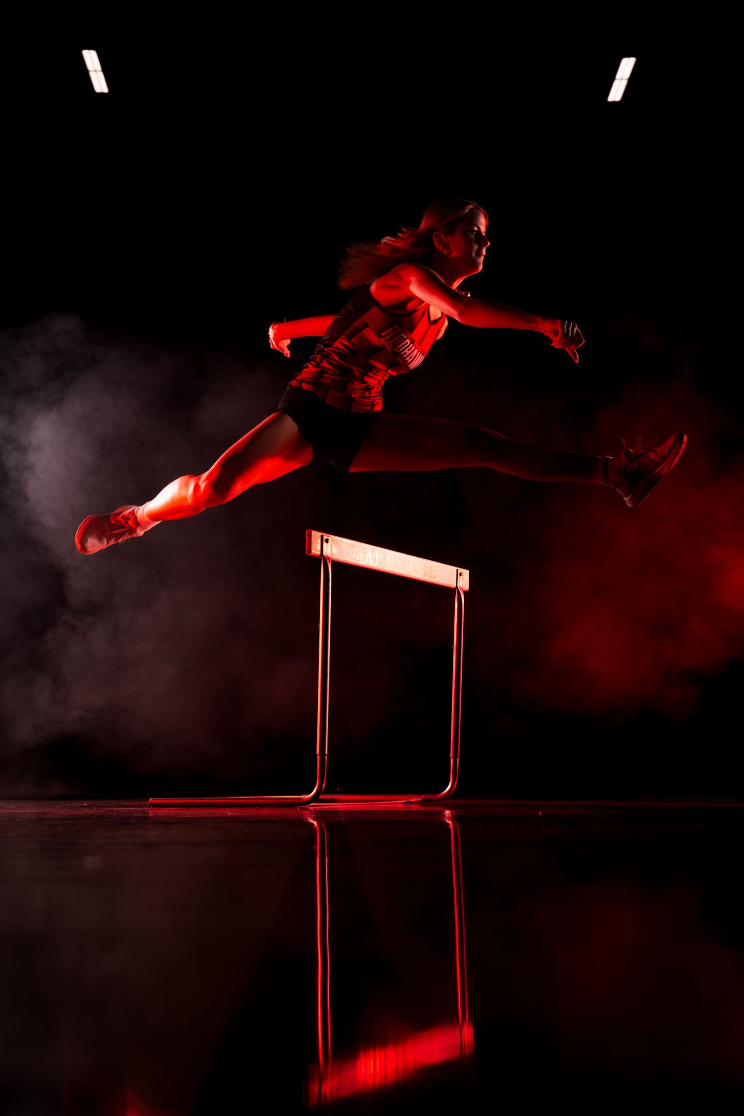 Silhouetted athlete jumping over hurdles against dramatic red lighting.
