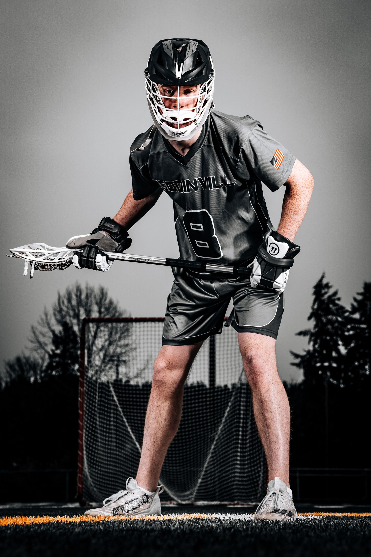 Lacrosse player in black uniform and protective gear crouches in defensive stance on field at dusk.