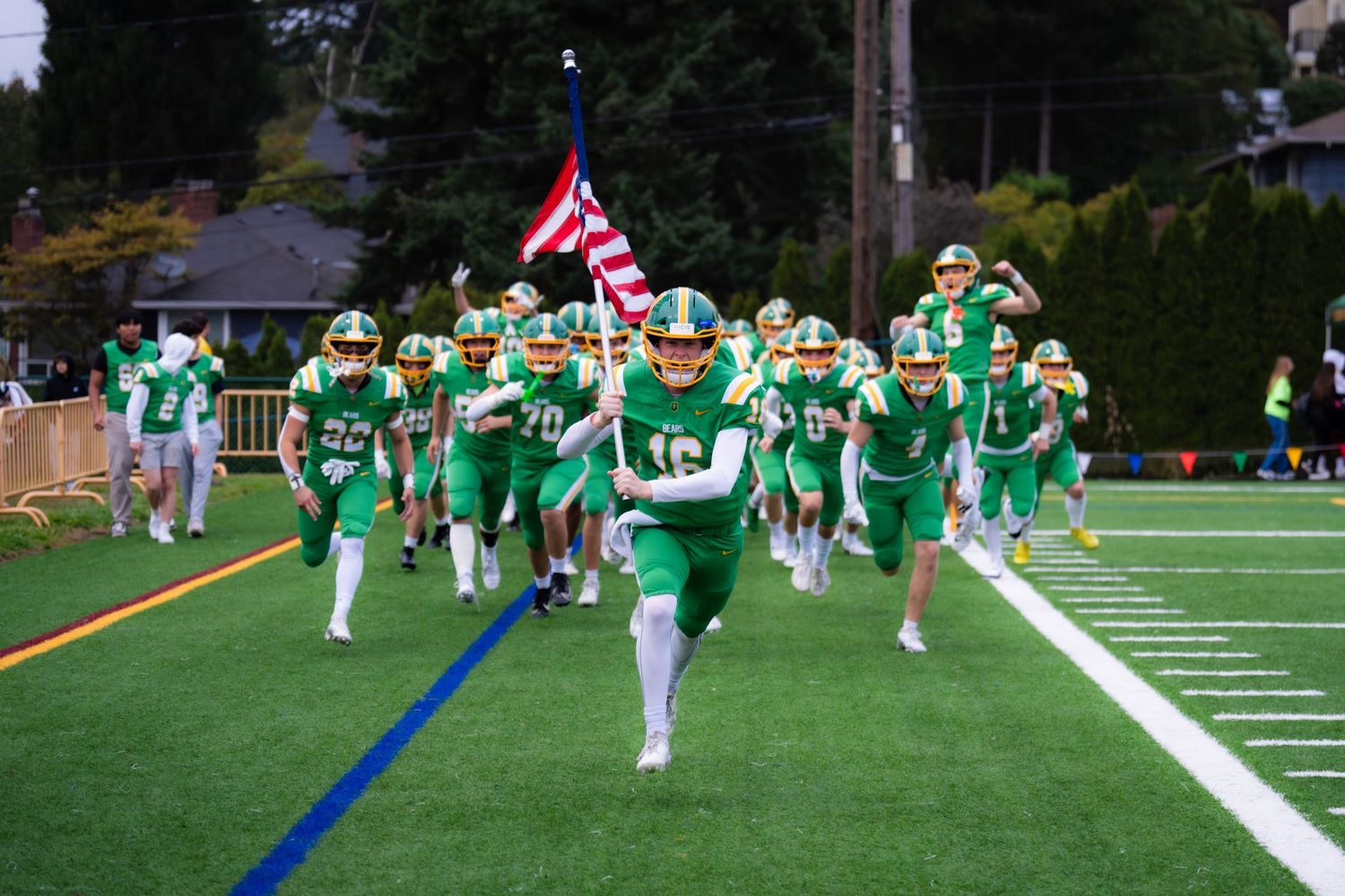 Football players in green and white uniforms practice formations on a turf field during team training session.