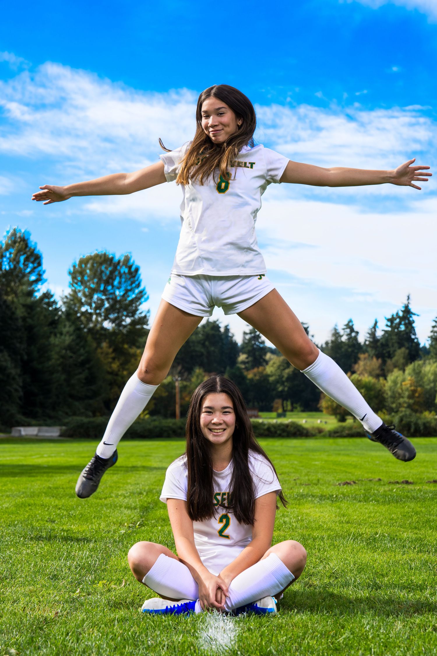 Young woman in white outfit performs playful yoga poses on sunny green grass field against blue sky.