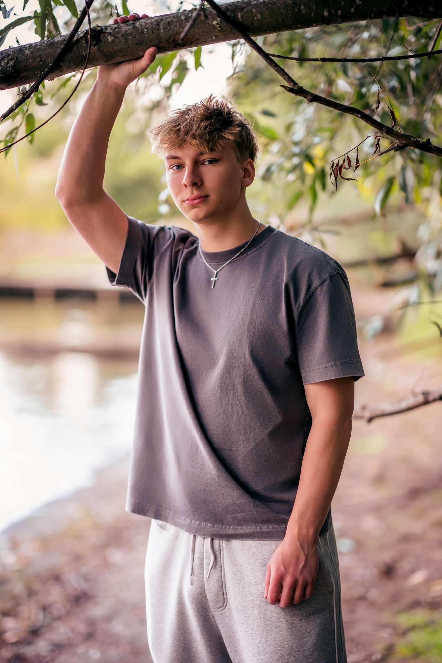A person in a gray t-shirt poses casually under tree branches near a lake.