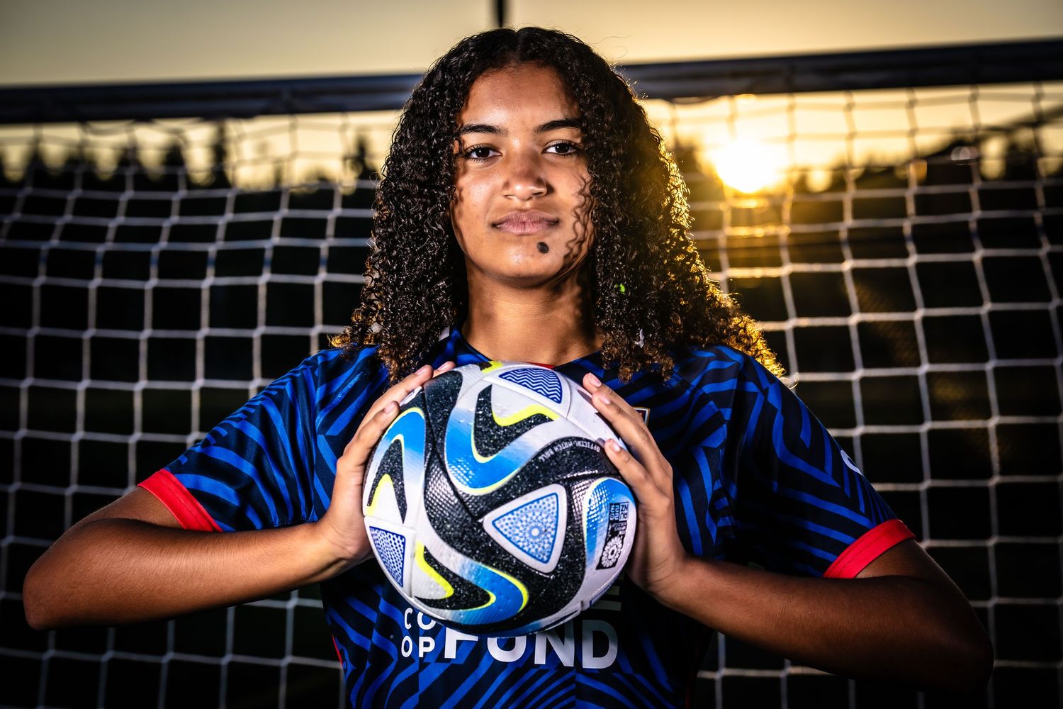Soccer player holds official FIFA match ball in front of goal net during golden hour sunset.