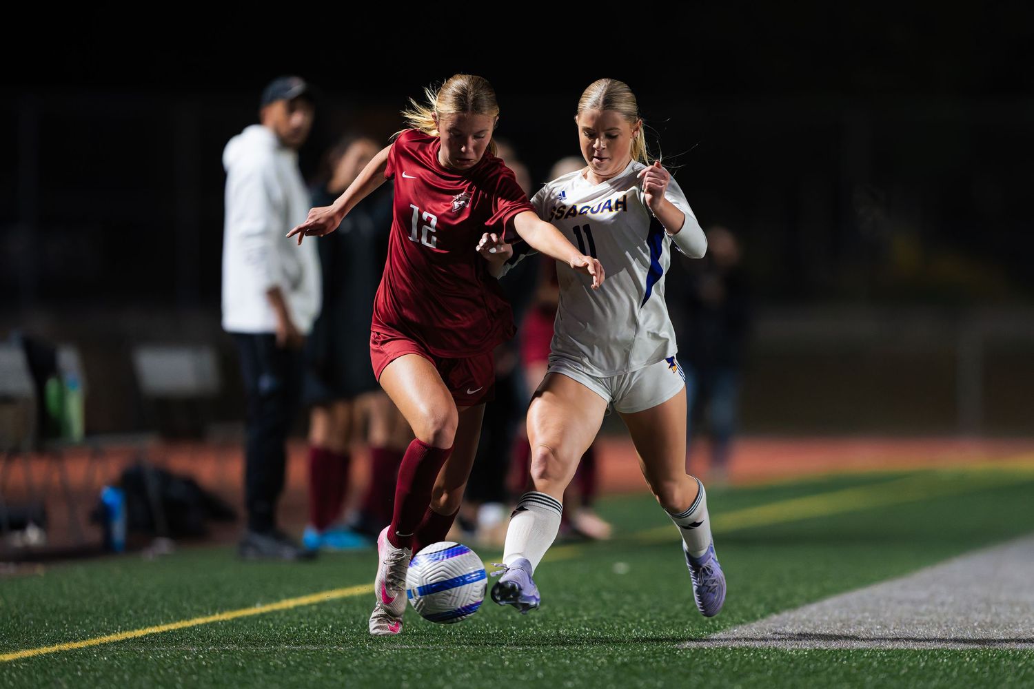 Two soccer players battle for possession of the ball during a nighttime match on an artificial turf field.