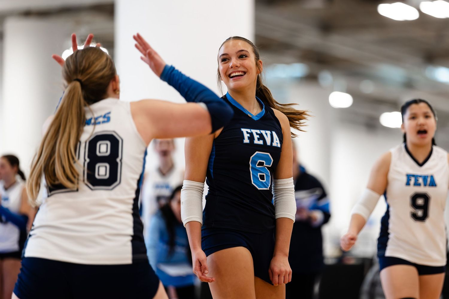 Volleyball players celebrate during an indoor match with one player wearing a black jersey raising her arms in excitement.