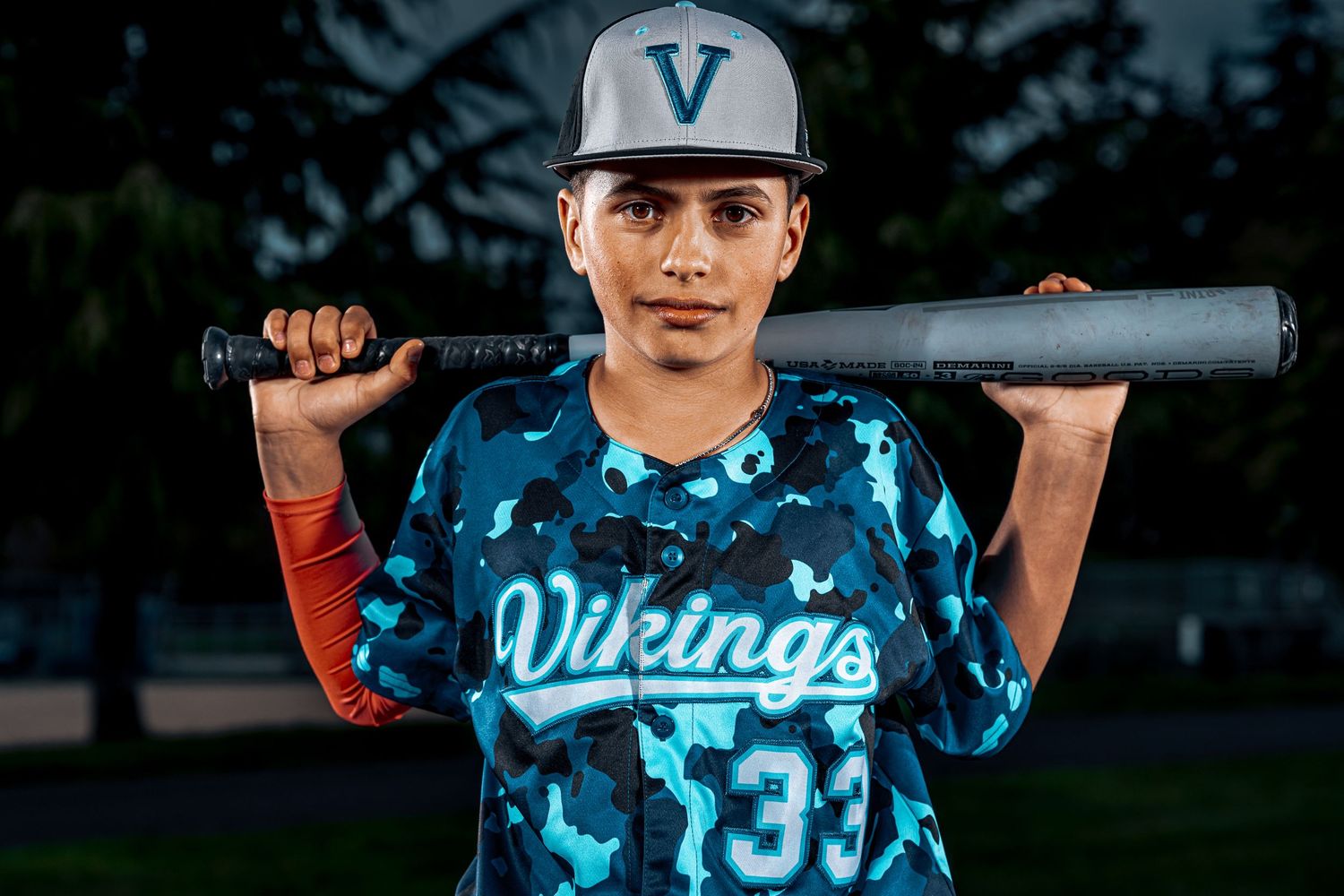 Baseball player in blue camouflage uniform holds bat during twilight game practice session.