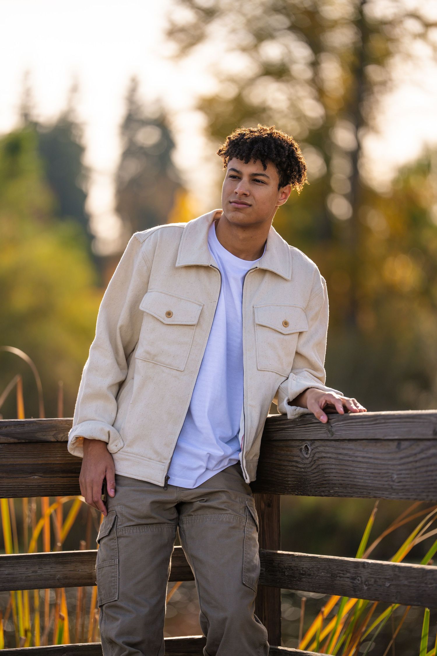 Person in beige jacket and white shirt sitting casually on wooden fence at sunset.