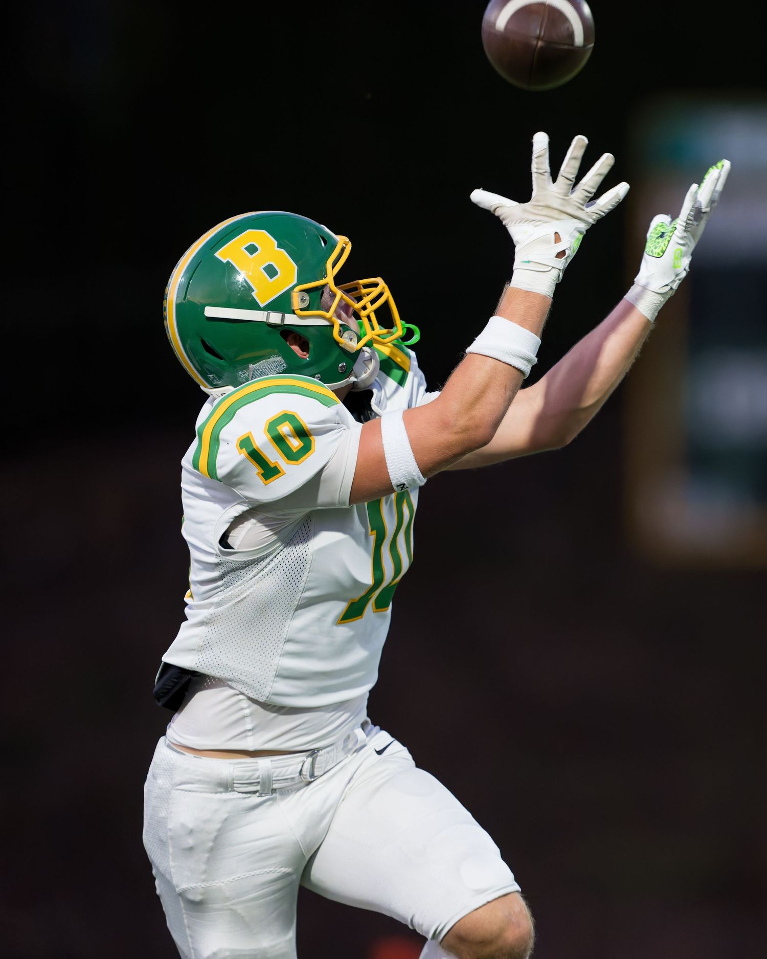 Football player in white uniform and green helmet leaps high to catch a pass during a game.