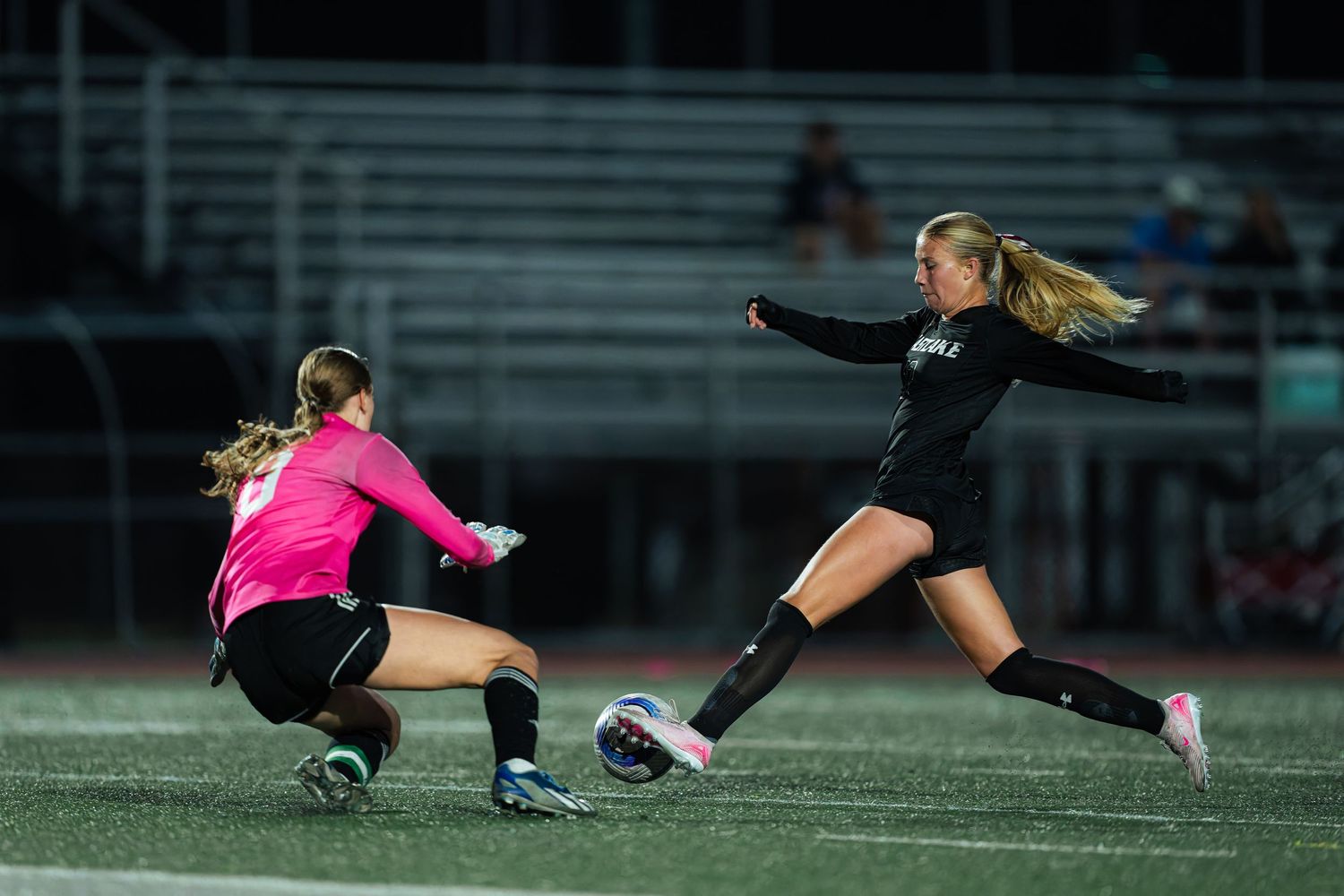 Soccer goalkeeper in pink jersey dives to make a save during a match on artificial turf field.