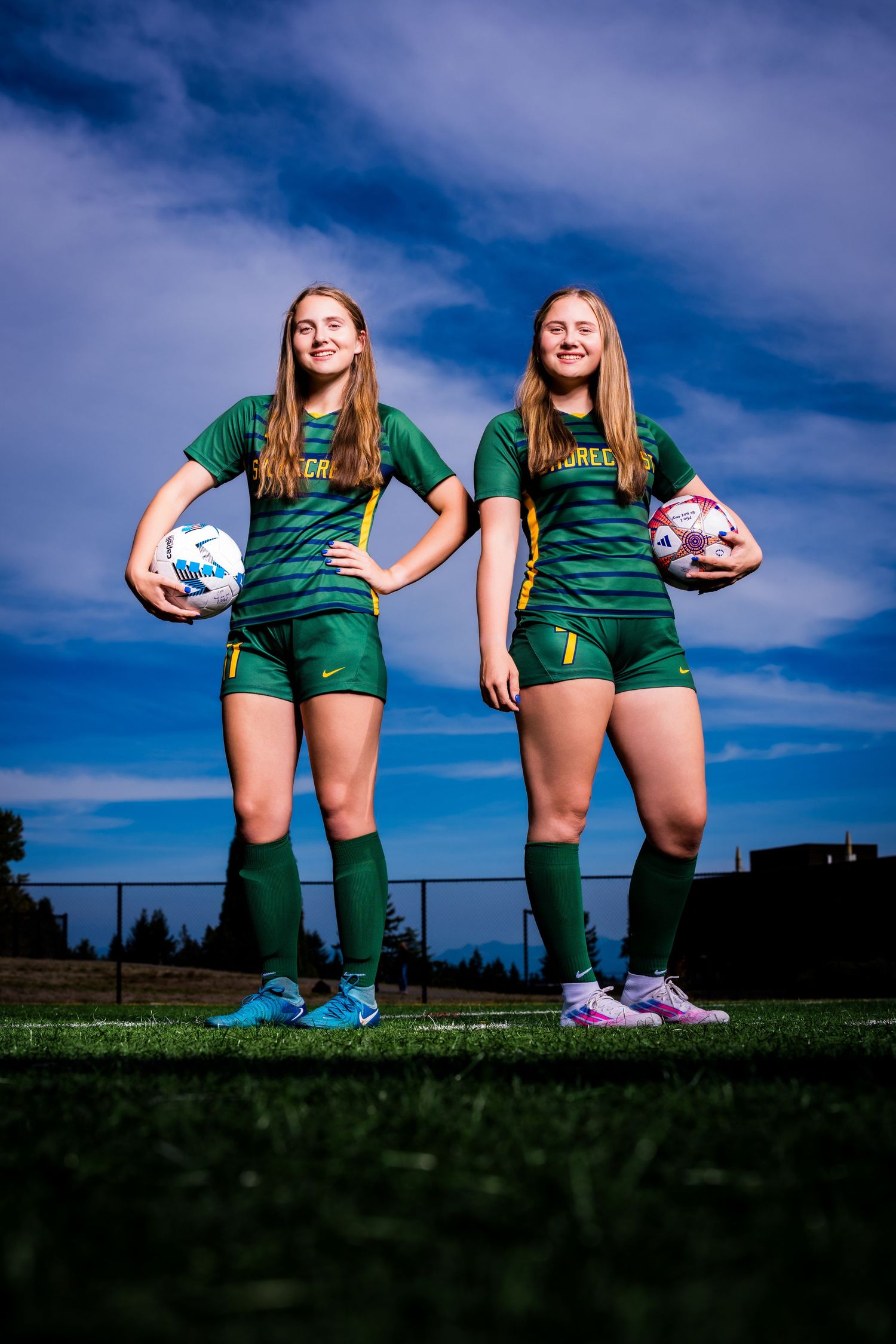 Soccer players in green uniforms stand together on field holding pink and white soccer balls against blue sky.