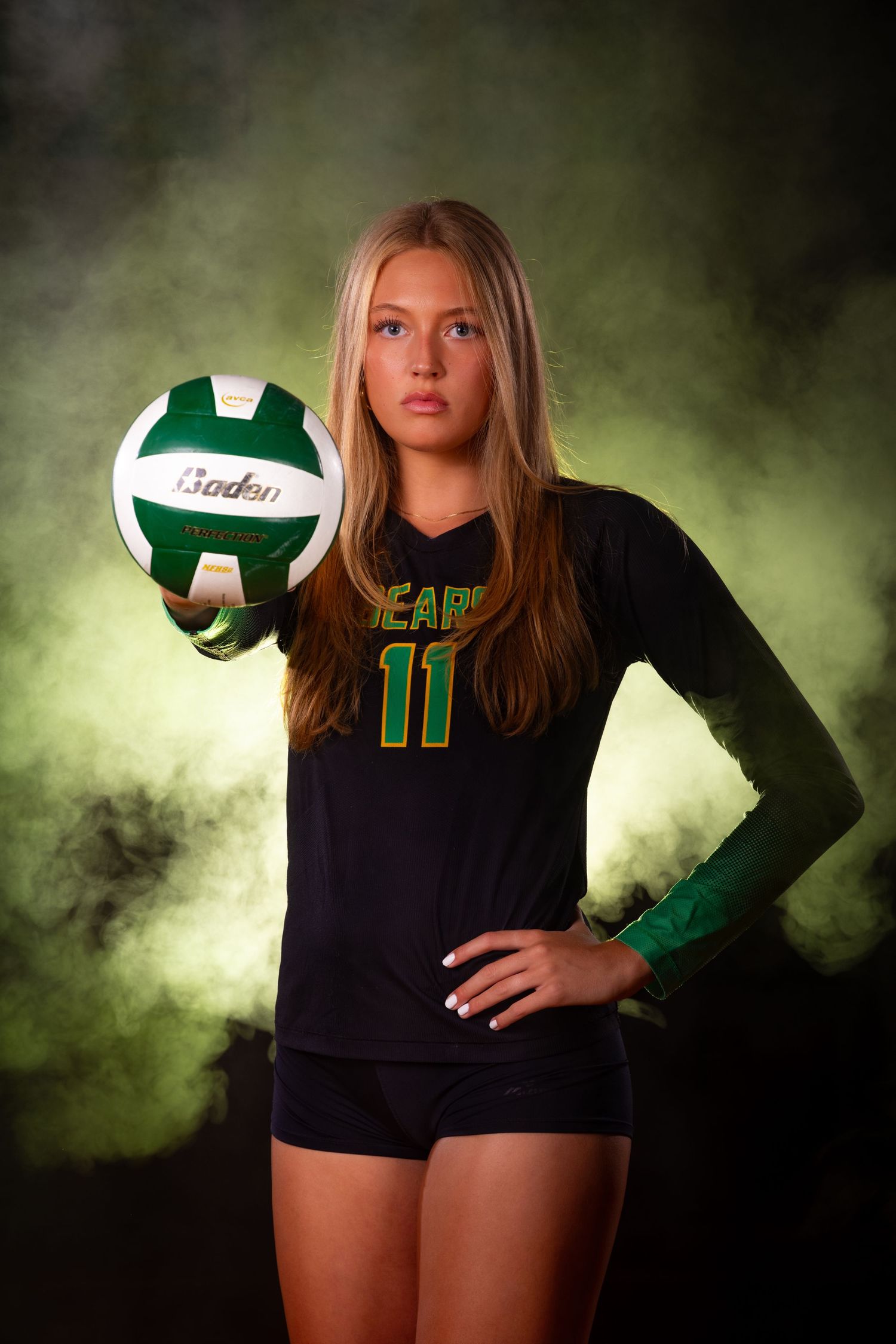 Volleyball player in dark uniform poses with ball against dramatic green lighting.