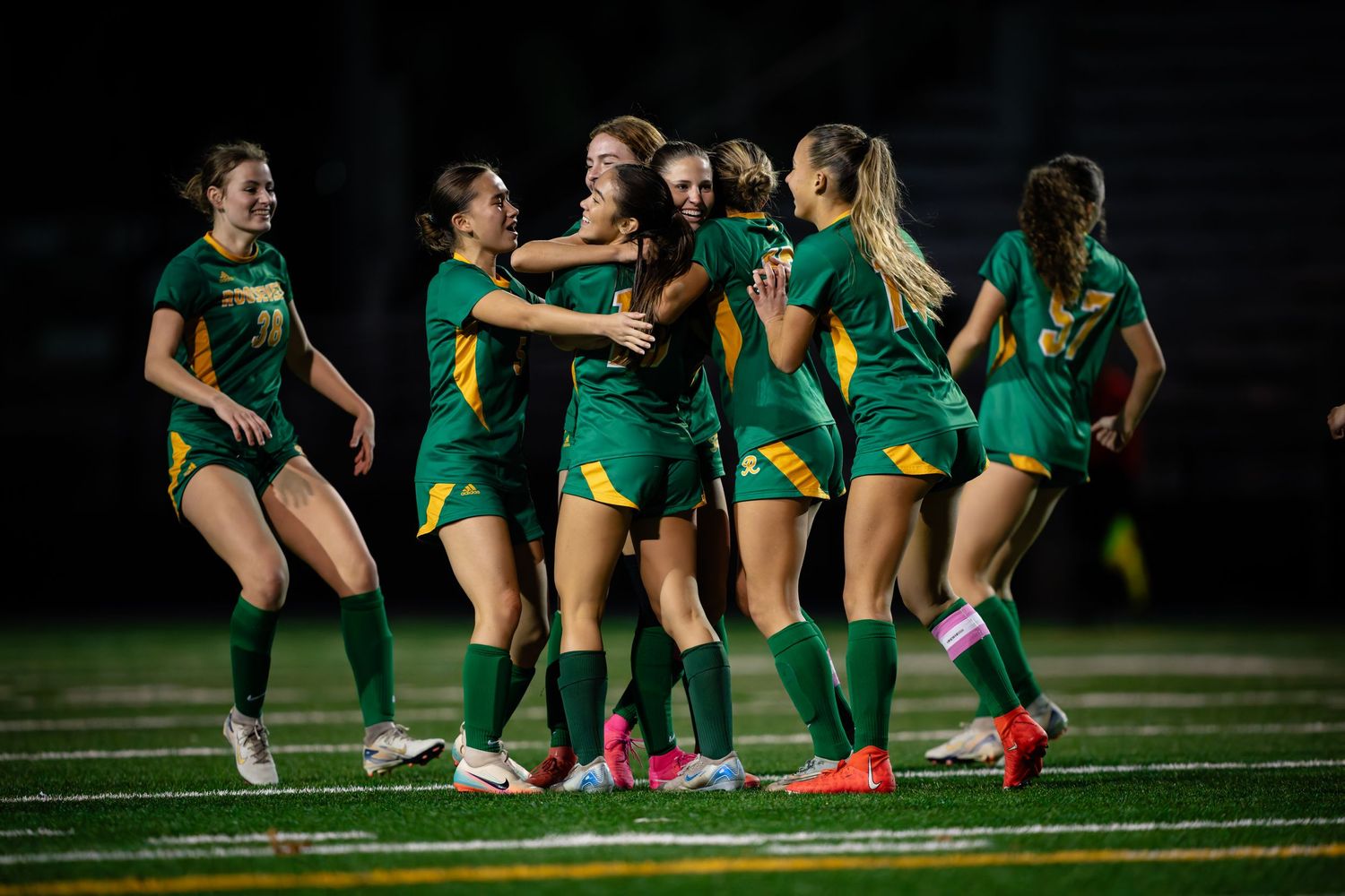 Female soccer players in green uniforms celebrate together on a field at night during a match.