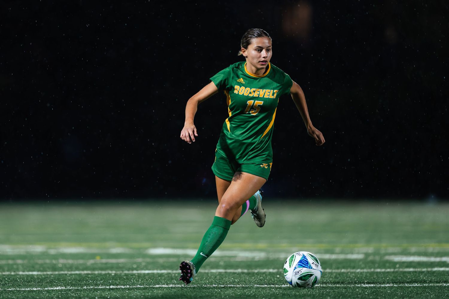 Soccer player in green uniform dribbles ball across field during nighttime match under stadium lights.