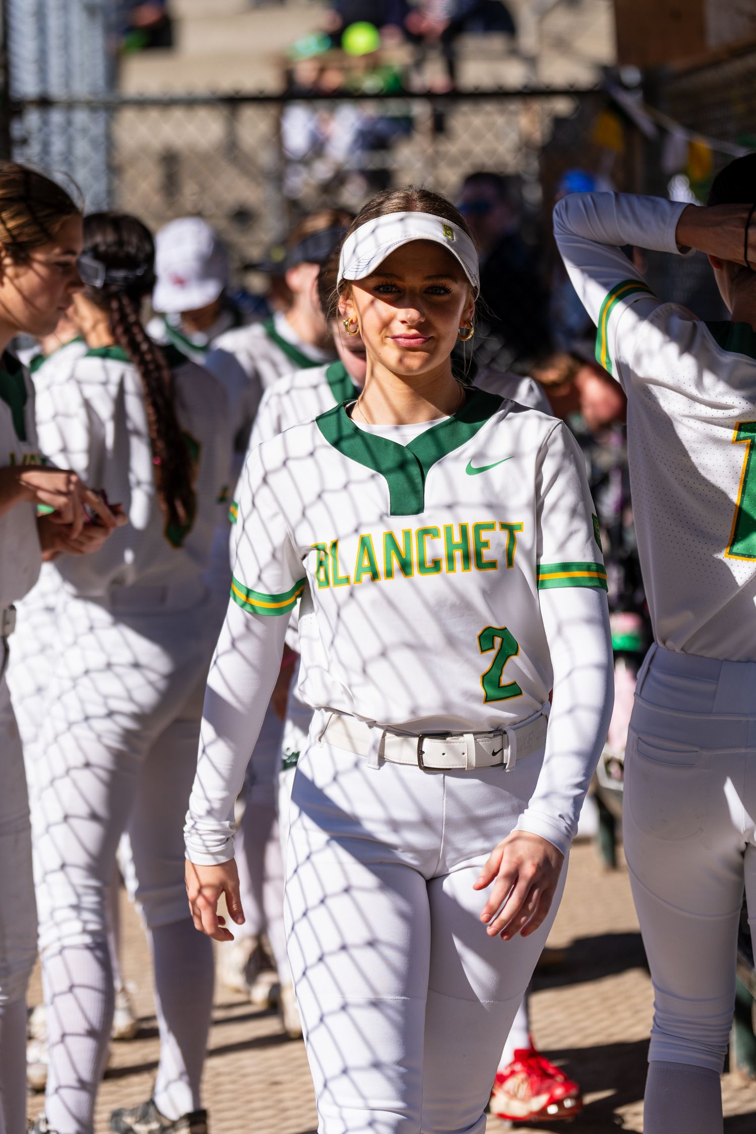 Softball player wearing white and green Blanchet uniform with number 2 stands in dugout during sunny game.
