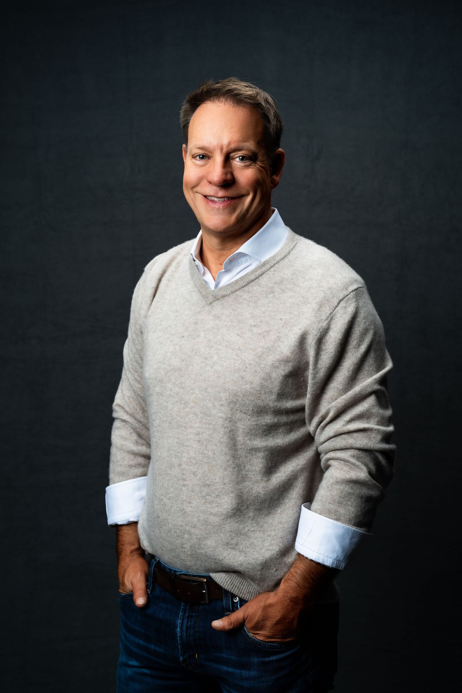 Professional studio portrait of someone wearing a gray v-neck sweater over light blue dress shirt with jeans against dark backdrop.