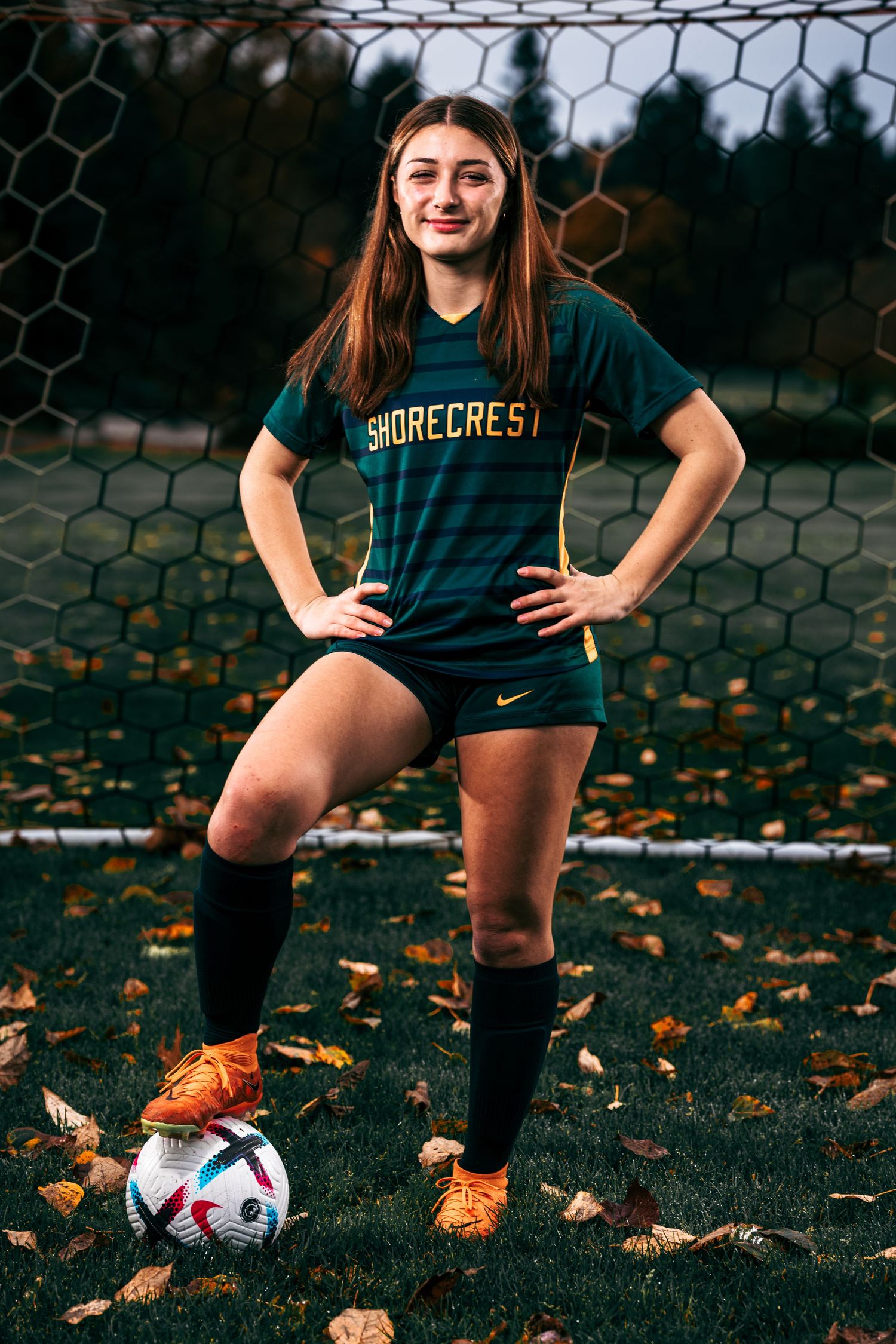Soccer player stands in front of goal net with autumn leaves scattered on grass field and ball at feet.