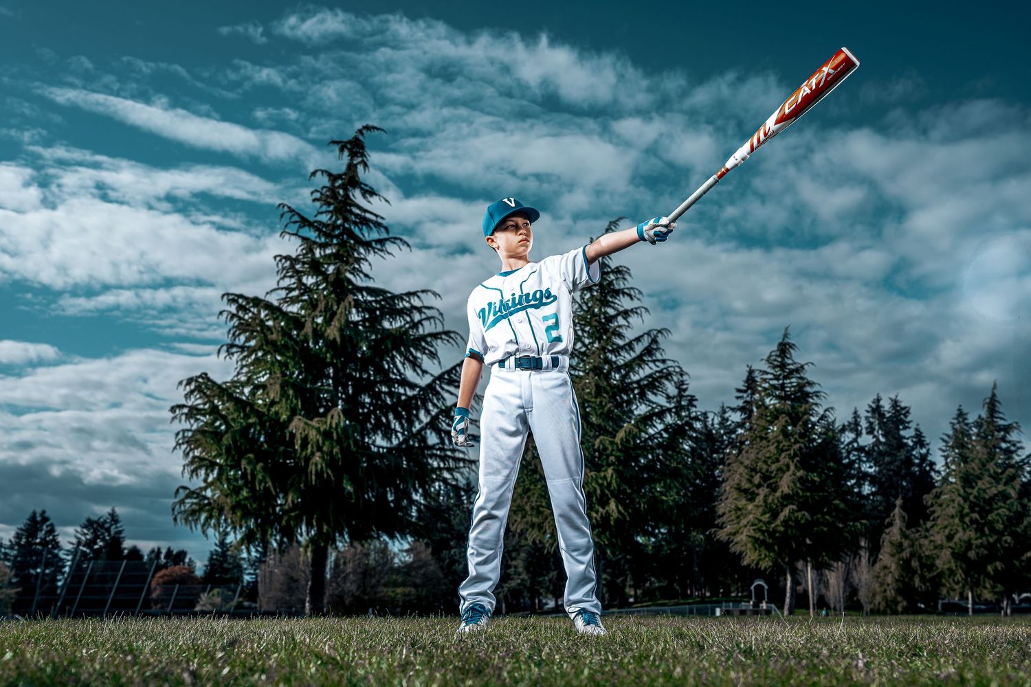 Baseball player in white uniform swings bat during outdoor practice session against dramatic cloudy sky.