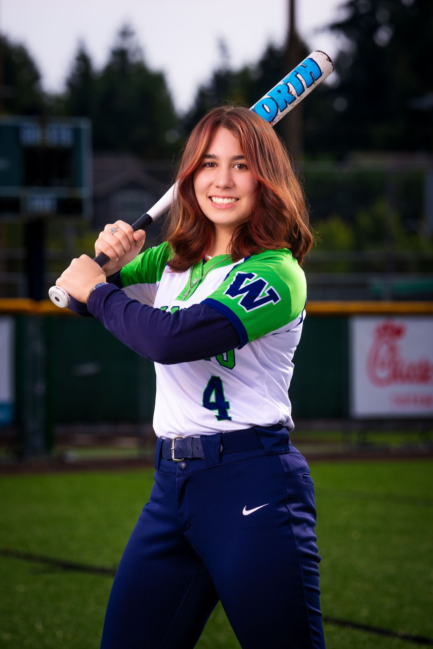 Softball player in blue and green uniform poses with bat on field.