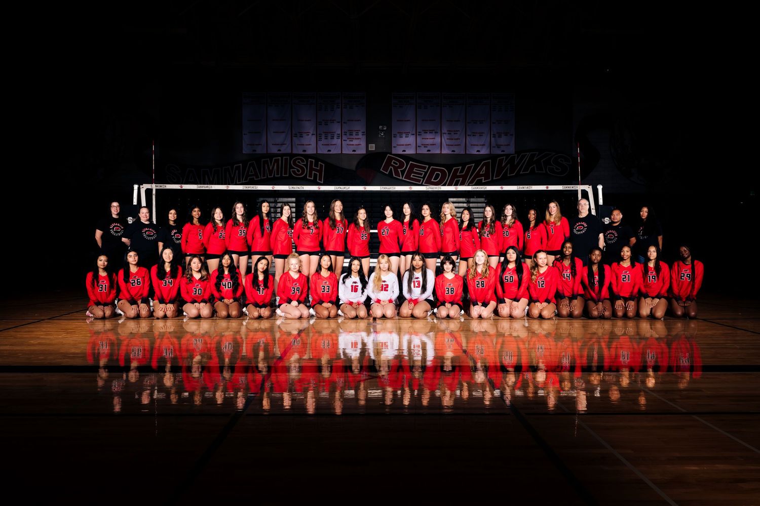 Sports team members in red uniforms stand in line formation on a dark stage during a formal presentation event.